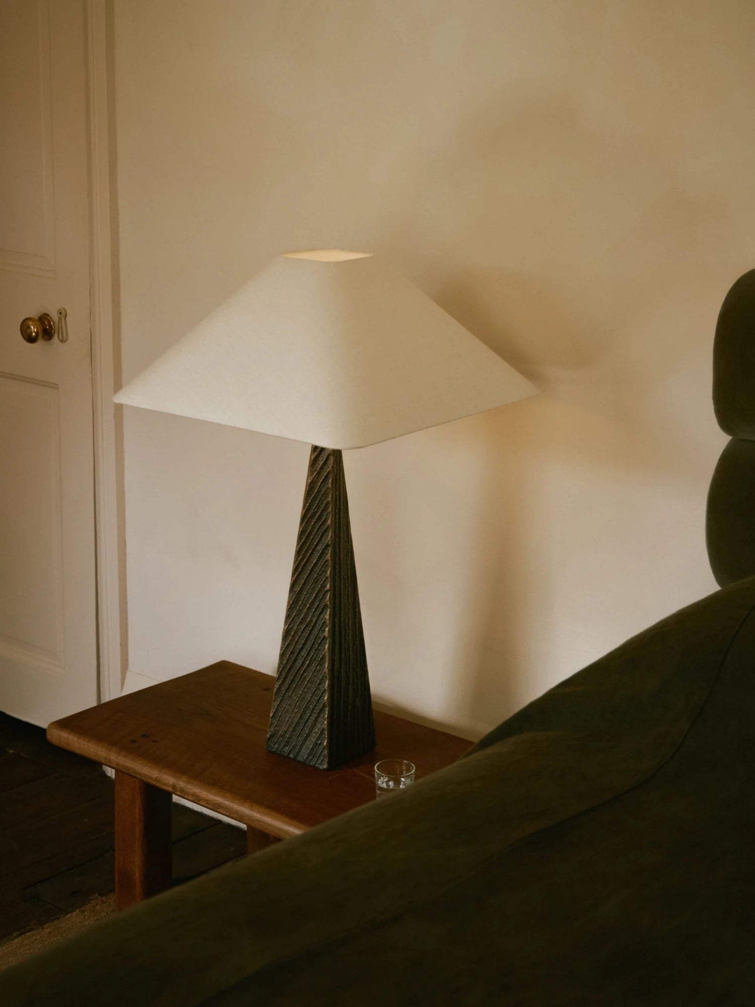 A minimalist Cubist Table Lamp with a textured, dark bronze or wood base is on a small wooden side table. The lamp has a large, square, off-white lampshade. A glass of water is also on the table, and the corner of a green velvet couch is visible on the right. A white door is in the background.
