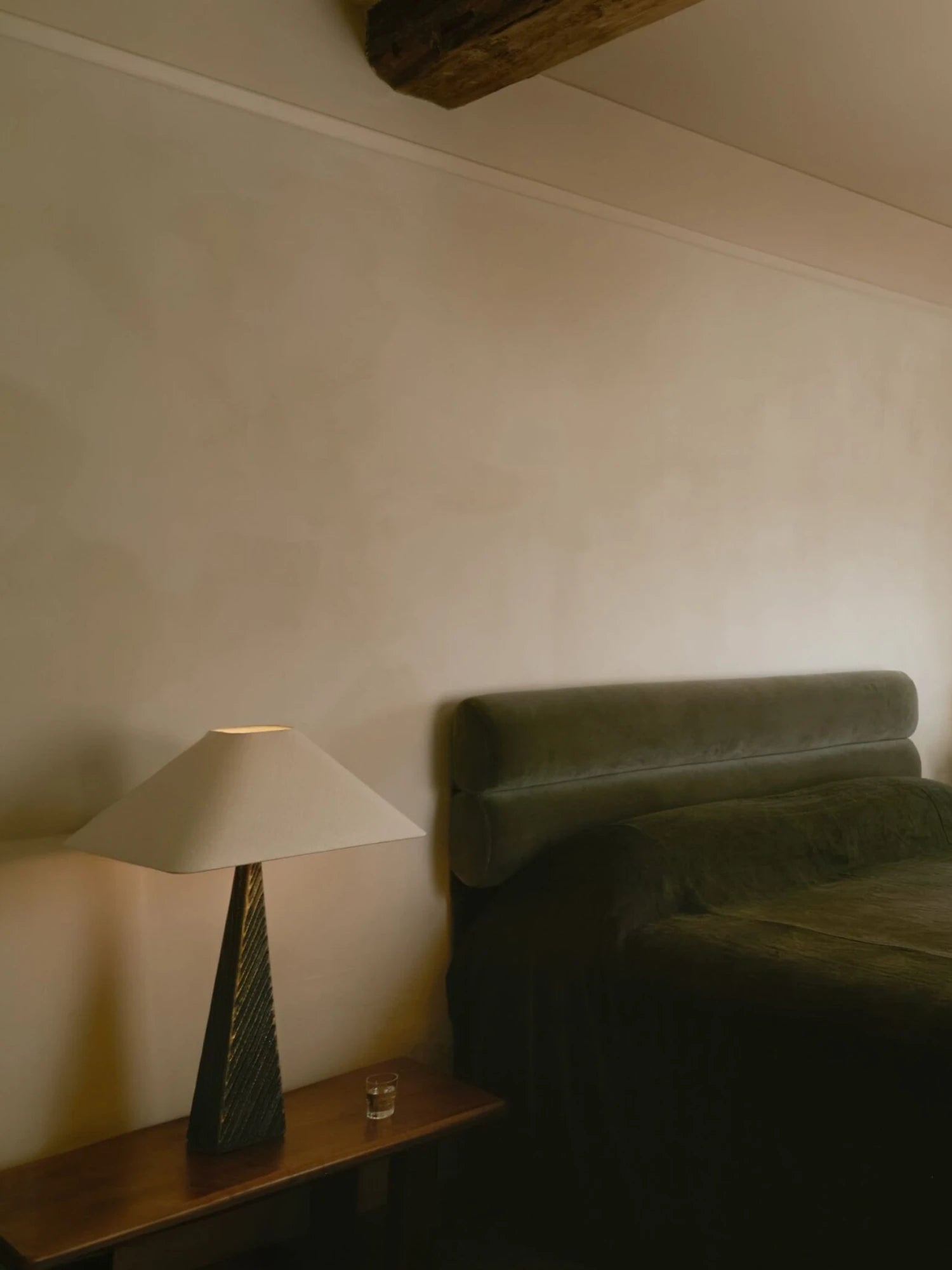 A bedroom scene with a low-light atmosphere features a Cubist Table Lamp on a wooden nightstand. The lamp has a dark, textured base and a wide, square, off-white lampshade that casts a warm glow. A bed with a plush, olive-green headboard and comforter is visible next to the nightstand. There is a small glass of water on the nightstand and a wooden beam on the ceiling.