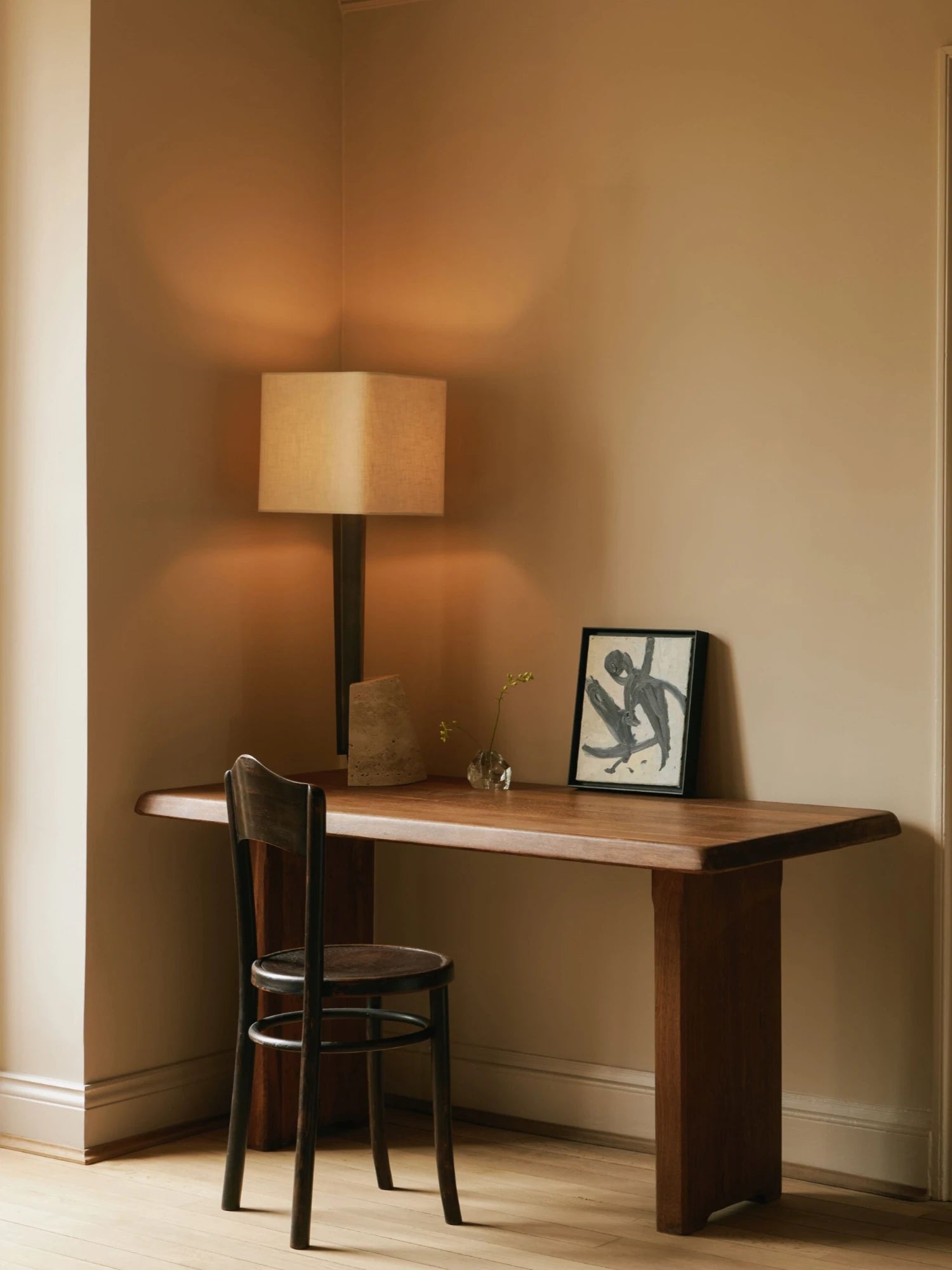 A wooden desk with two thick legs is positioned in the corner of a room with warm, neutral-colored walls. A tall Roc Table Lamp with a square, textured lampshade is on the desk, casting a warm glow on the wall. A small, clear glass vase with a single stem, a small rock-like sculpture, and a framed abstract art print are also on the desk. A dark wood chair is in front of the desk, facing away from the viewer.