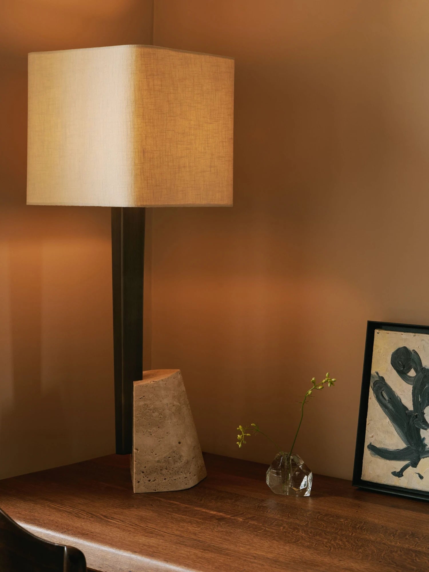 A tall Roc Table Lamp with a rectangular, off-white lampshade is on a wooden desk. The lamp's base is a dark, sleek, and minimalist design. Next to it, there is a small, clear glass vase holding a thin green stem with a few flowers, an abstract concrete or stone sculpture, and a framed piece of abstract art with black and yellow shapes. The walls are a warm, earthy tone.