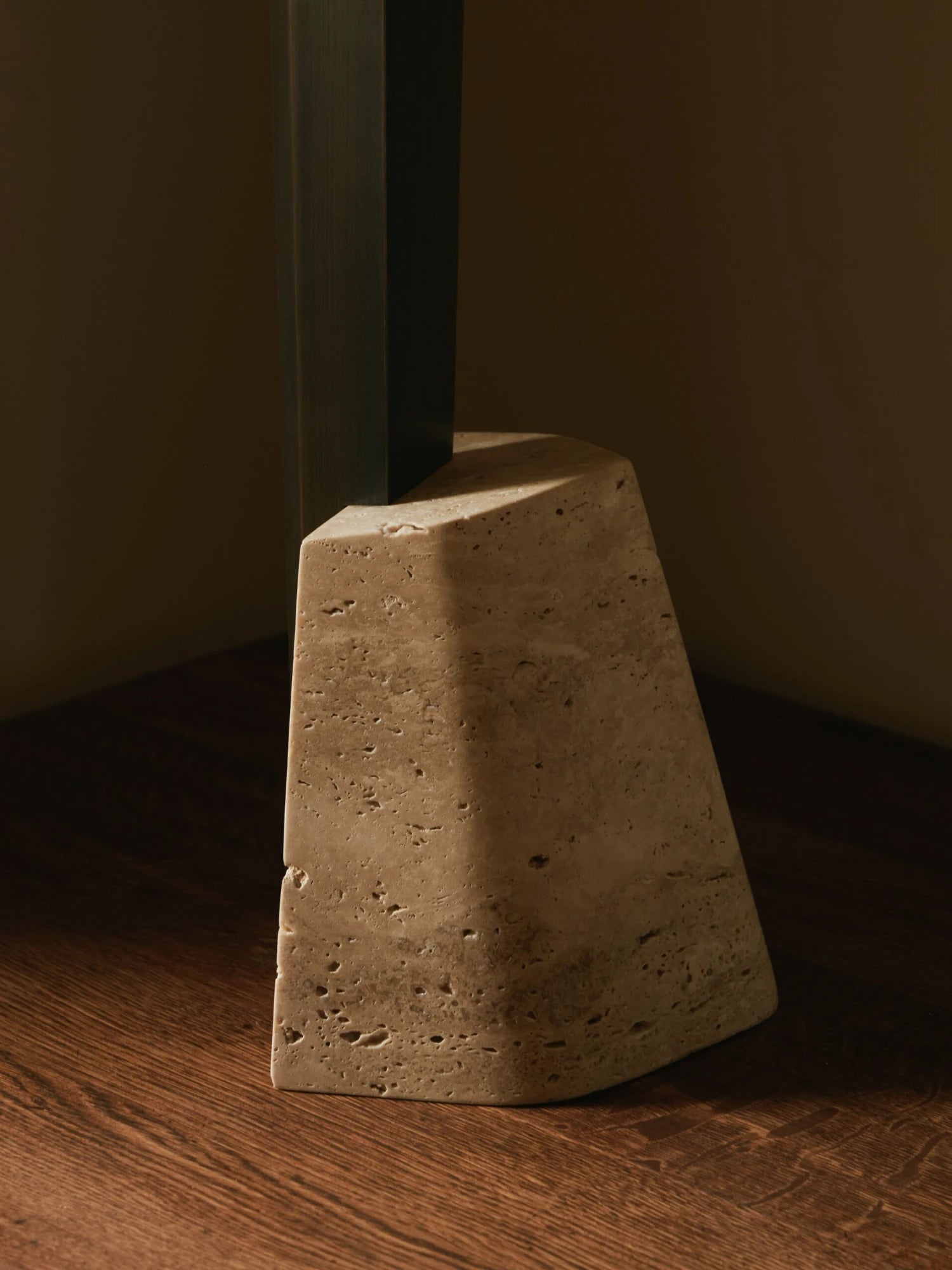 A close-up shot of the base of a Roc Table Lamp. The base is made of a dark, sleek material and is set into a separate, chunky, wedge-shaped object made of porous concrete or stone. The lamp and the stone are on a wooden surface in a corner. Sunlight casts a shadow on the objects, highlighting the textures.