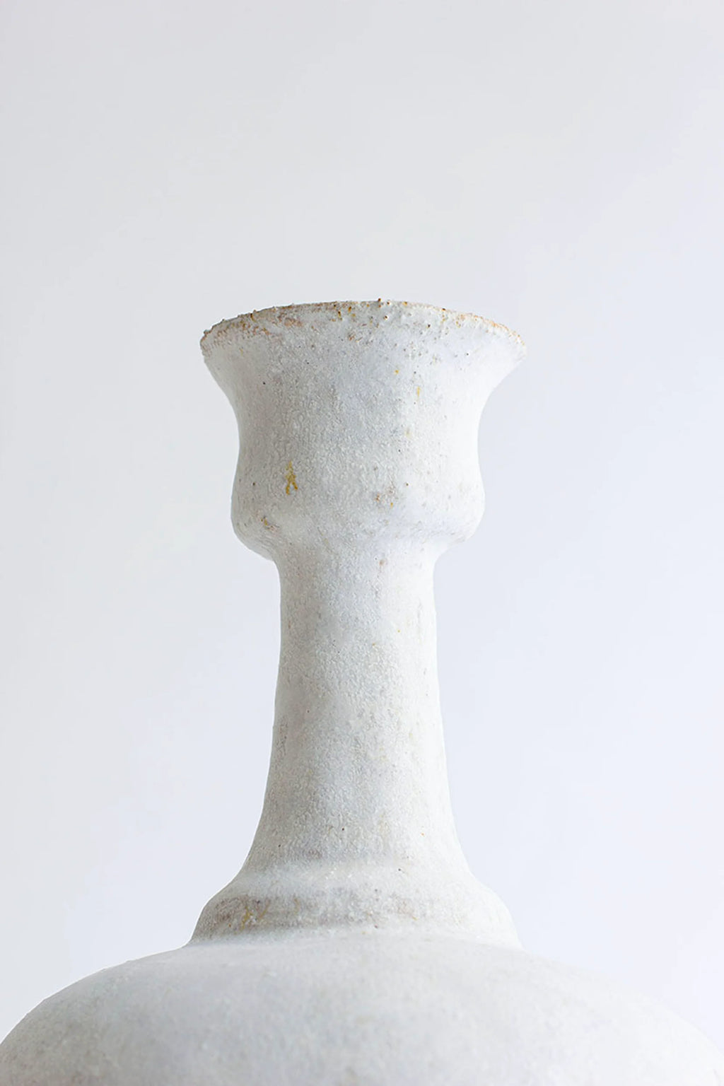 Canoa Lab Arq 005 Blanco Hueso Vase, a handcrafted ceramic vase in a warm blanco hueso (bone white) finish, featuring a unique, highly textured porous surface reminiscent of volcanic rock or coarse sand. Its sculptural, organic form includes a slender neck with a flared, goblet-like mouth, above a rounded body. This modern minimalist stoneware piece is perfect for organic modern shelf styling, adding rustic elegance to living rooms or entryways.
