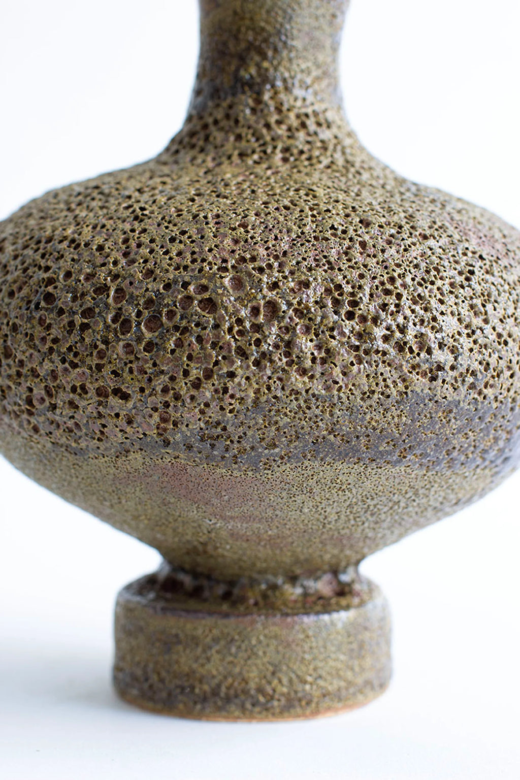 Canoa Lab Arq 006 Musgo Vase, a handcrafted stoneware vase featuring a unique, highly textured porous surface resembling volcanic rock in earthy moss green and deep brown tones. This organic modern ceramic vase boasts a bulbous body, a slender neck, and a sturdy cylindrical base, showcasing a raw, wabi-sabi aesthetic with its rich, natural color variations. Ideal for rustic shelf styling, a natural accent in contemporary living rooms, or as an organic centerpiece for brutalist or japandi-inspired interiors.