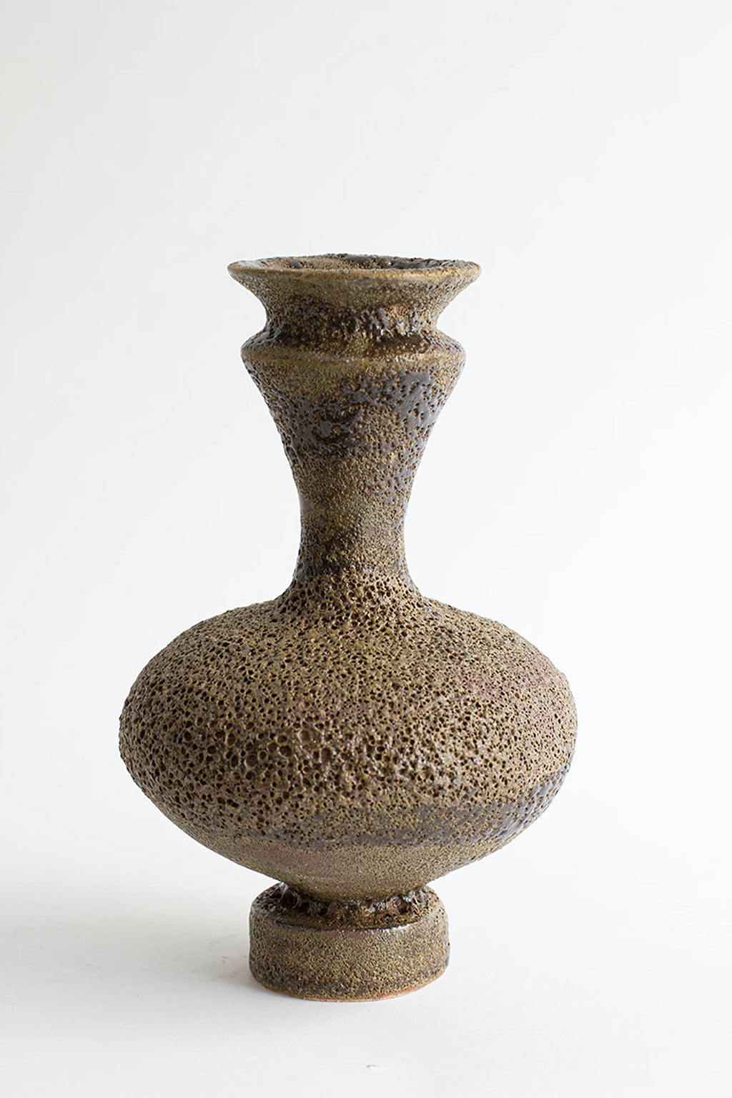 Canoa Lab Arq 006 Musgo Vase, a handcrafted stoneware vase featuring a distinctive porous, volcanic texture in rich moss green and earthy brown tones with dark brown glazed accents. Its organic, sculptural silhouette includes a bulbous body, a long tapering neck, and a flared rim, all resting on a sturdy cylindrical base. This unique decorative ceramic vase is perfect for adding a rustic, wabi-sabi, or organic modern touch to living room shelves, entryways, or contemporary interior styling.