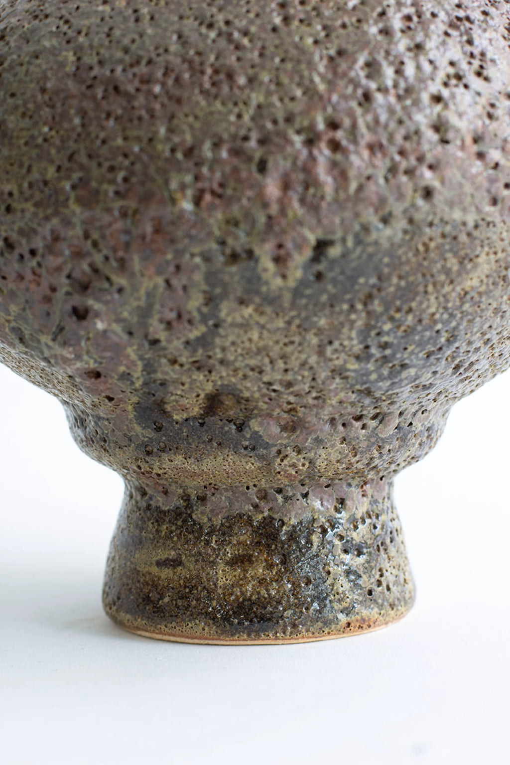 Canoa Lab Arq 007 Musgo Vase, a detailed view of the rustic, handcrafted ceramic vase's base, showcasing its textured, porous surface in earthy brown and green tones, on a white background.