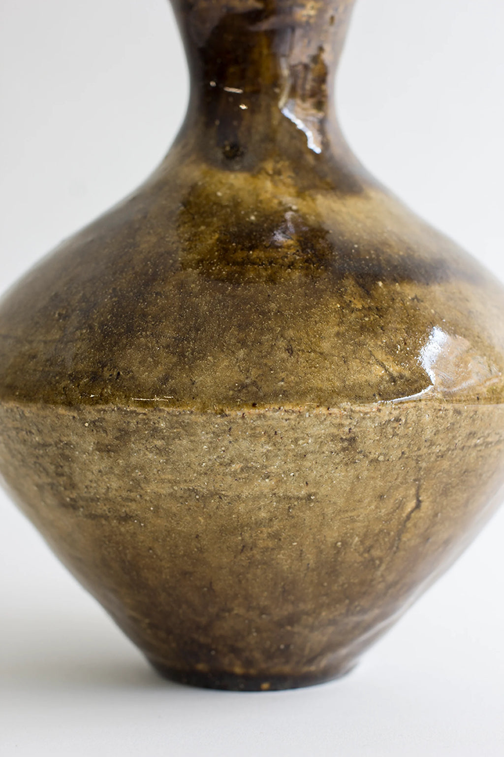Canoa Lab Arq 008 Vase, a handcrafted organic modern ceramic vase featuring a distinctive two-tone finish. The upper half showcases a rich, dark brown and olive green reactive glaze with subtle drips, while the lower body transitions to a rough, unglazed raw stoneware texture in earthy sand tones, creating an artisanal contrast. Its elegant, amphora-like silhouette is ideal for wabi-sabi shelf styling or as a rustic centerpiece in contemporary living rooms and natural-inspired spaces.