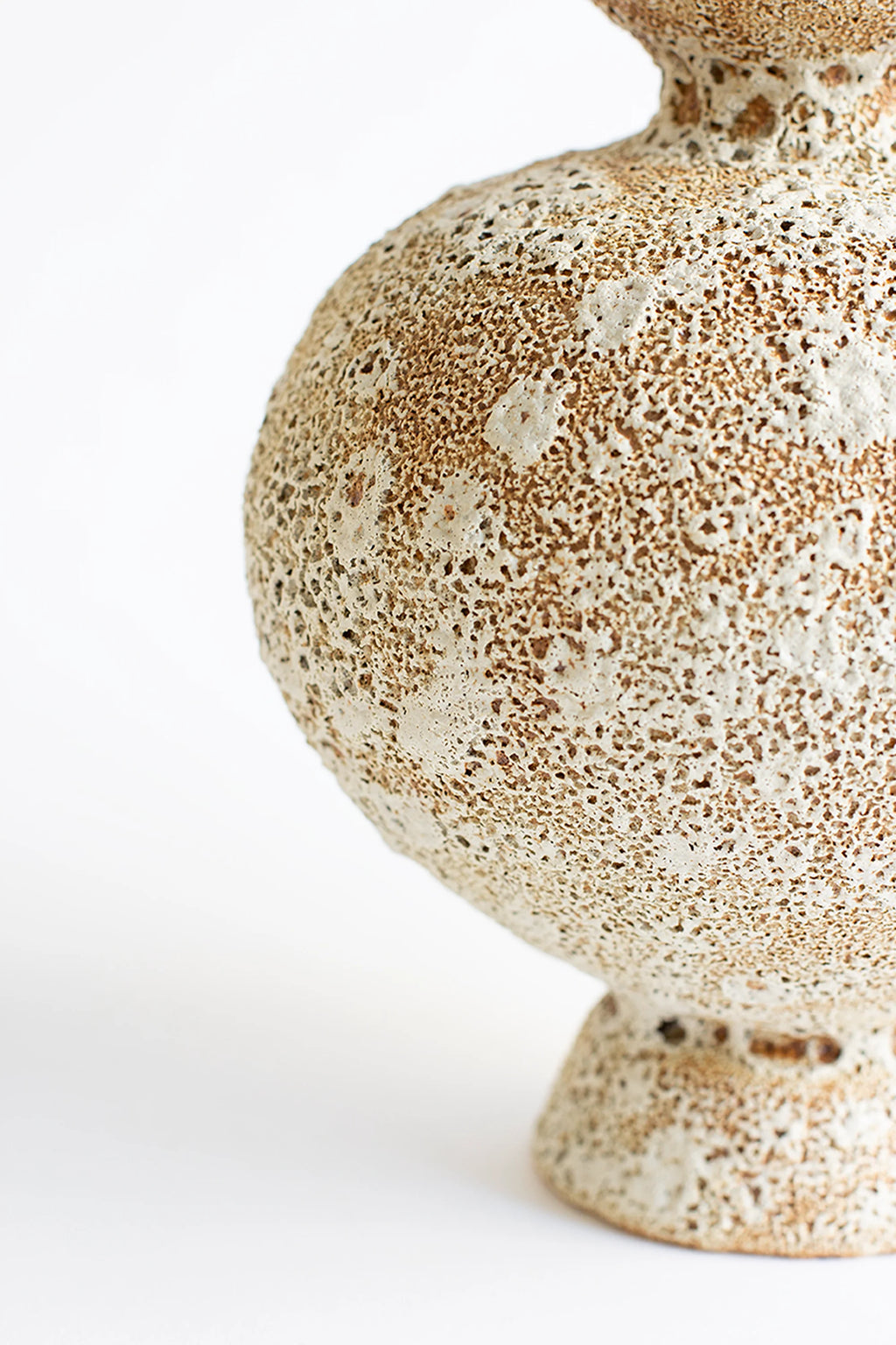 Canoa Lab Arq 013 Vase, a detailed side view of the lower, rounded section of the modern ceramic vase. The textured surface displays a unique pattern of raised white speckles against a warm, sandy beige, emphasizing its artisanal and organic design. Displayed on a white background.