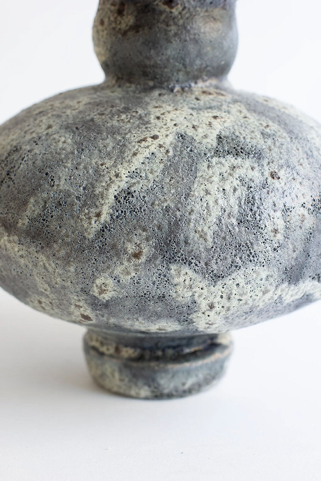 Canoa Lab Cálpide Gecko Vase, a close-up of the vase's lower body and base, revealing the rich, textured gray and white glaze with subtle brown speckles. The handcrafted ceramic detail is prominent, displayed on a white background.