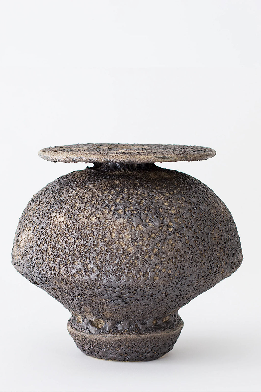 Canoa Lab Isolated n.18 Vase, a handcrafted ceramic vase with a dark charcoal and earthy brown volcanic texture, featuring a distinctive porous finish and a sculptural, bulbous body. Its unique organic form is topped with a flat, disc-like rim, evoking a brutalist or wabi-sabi aesthetic. This artisan stoneware piece is perfect for organic modern shelf styling or as a statement decorative object in contemporary minimalist living rooms.