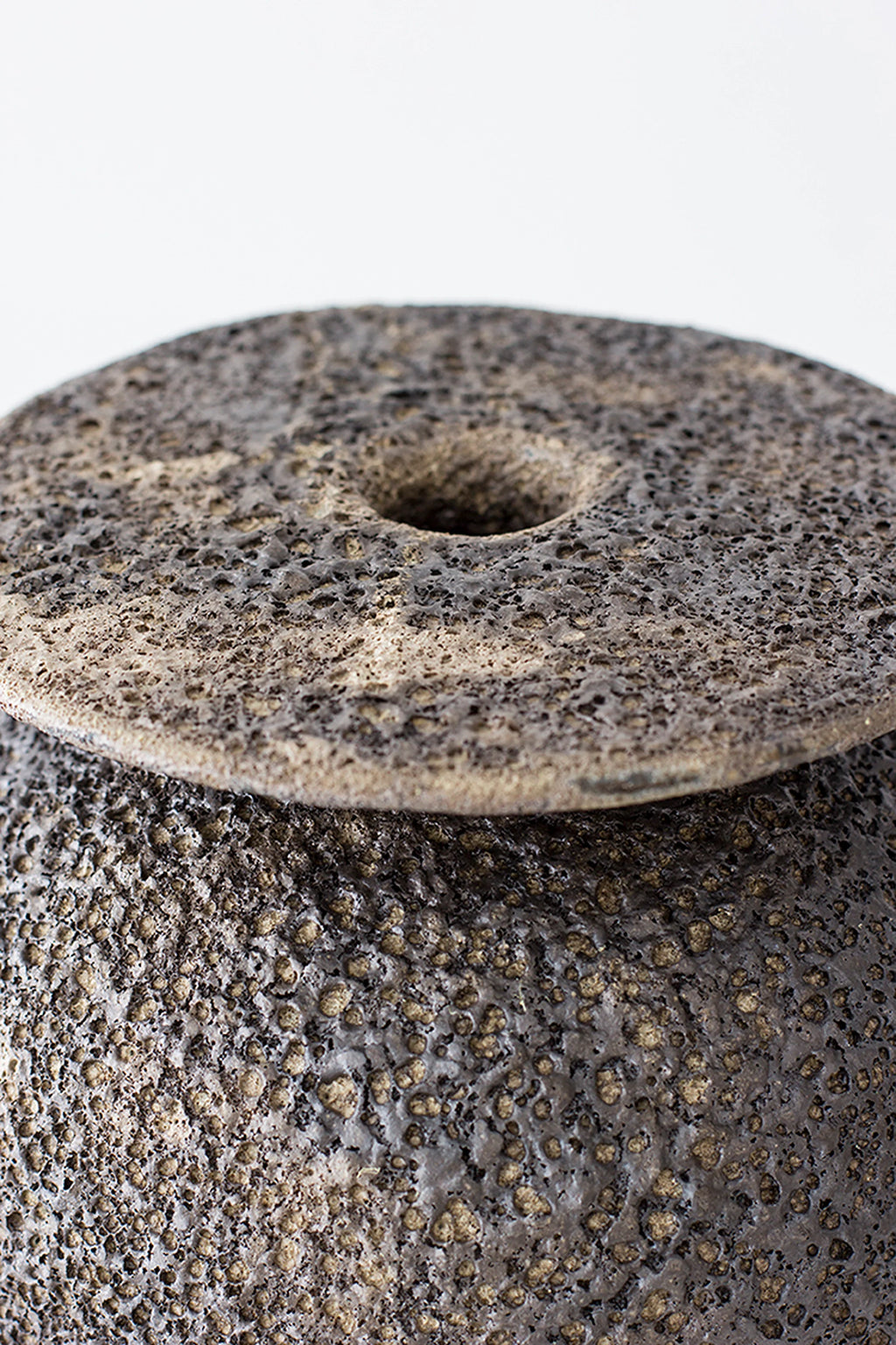 Canoa Lab Isolated n.18 Vase, a dark grey handcrafted ceramic vase featuring an intensely textured, porous volcanic finish and a unique disc-like top with a central opening. This artisanal stoneware piece showcases an organic modern aesthetic with rustic charm. Perfect for brutalist or japandi shelf styling in contemporary living spaces.