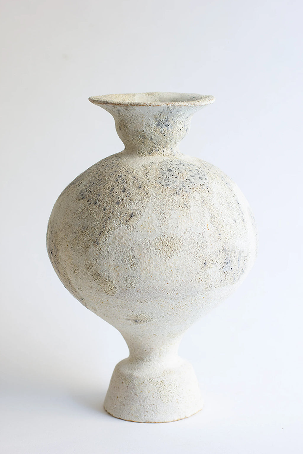 Canoa Lab Lekytho Cárabo Vase, a full, elegant view of the unique, bulbous ceramic vase with a narrow neck and flared rim, featuring a textured, off-white glaze with subtle dark speckles, standing on a white background.