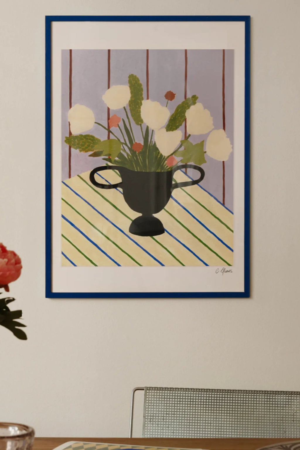 The Poster Club Flowers on Striped Cloth, a vibrant modern still life art print depicting cream-colored flowers and green foliage arranged in a black vase, set atop a yellow and cream tablecloth with bold green and blue diagonal stripes against a soft lilac background. This playful floral poster is presented in a vivid royal blue frame with a clean white mat, making it ideal as colorful wall art for contemporary living rooms, dining areas, or a cheerful accent in modern kitchens.