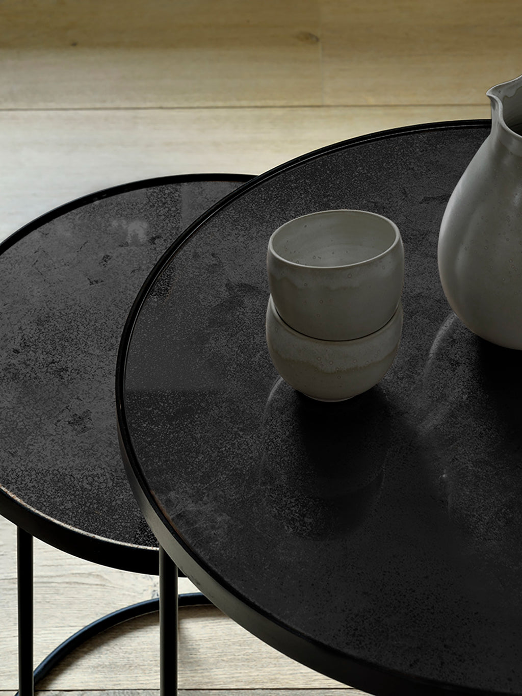 Ethnicraft Nesting Coffee Table Set featuring two charcoal-finished round tables with a textured, distressed surface. Styled with ceramic cups and a vase on a light wooden floor.