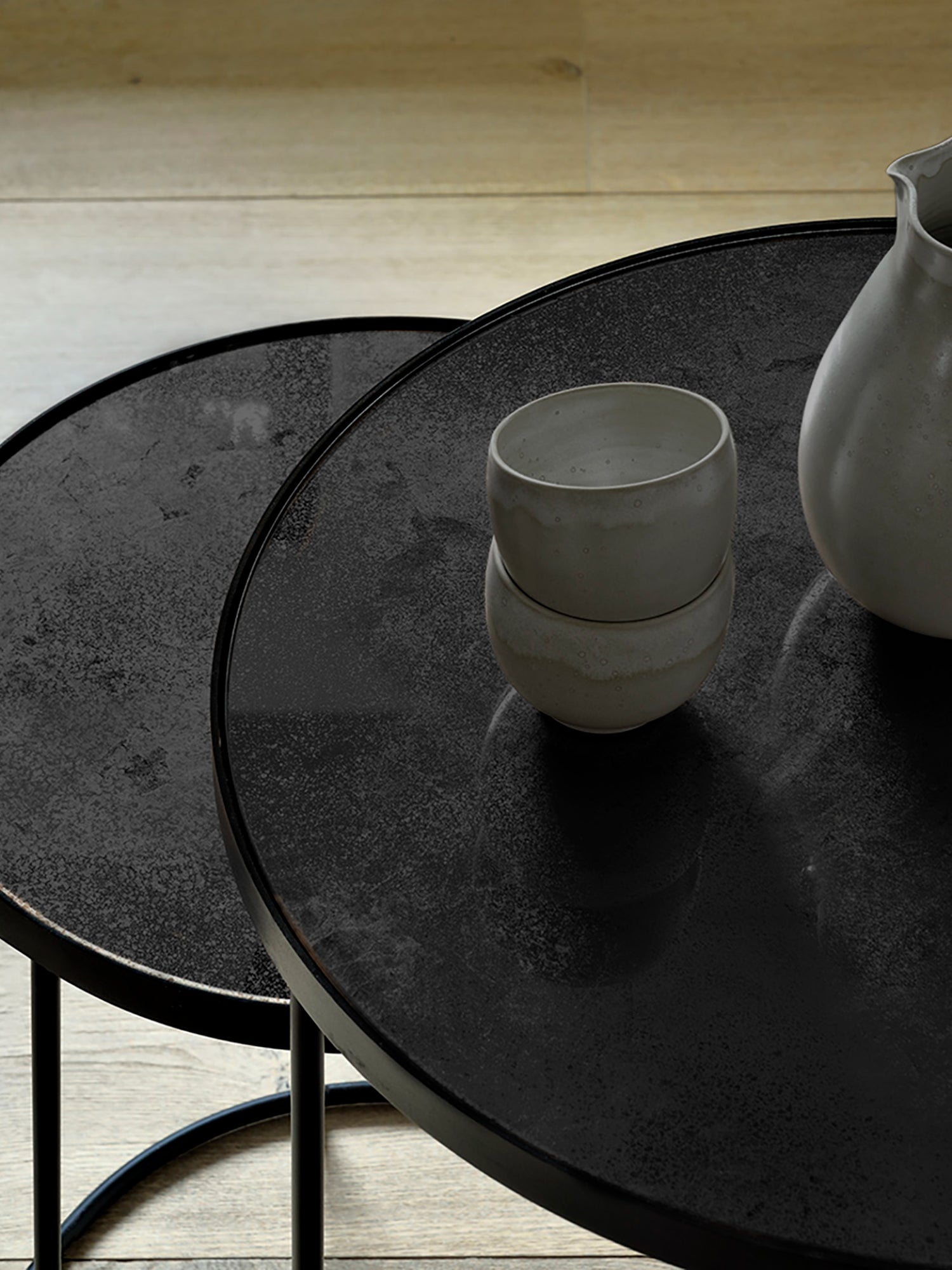Ethnicraft Nesting Coffee Table Set featuring two charcoal-finished round tables with a textured, distressed surface. Styled with ceramic cups and a vase on a light wooden floor.
