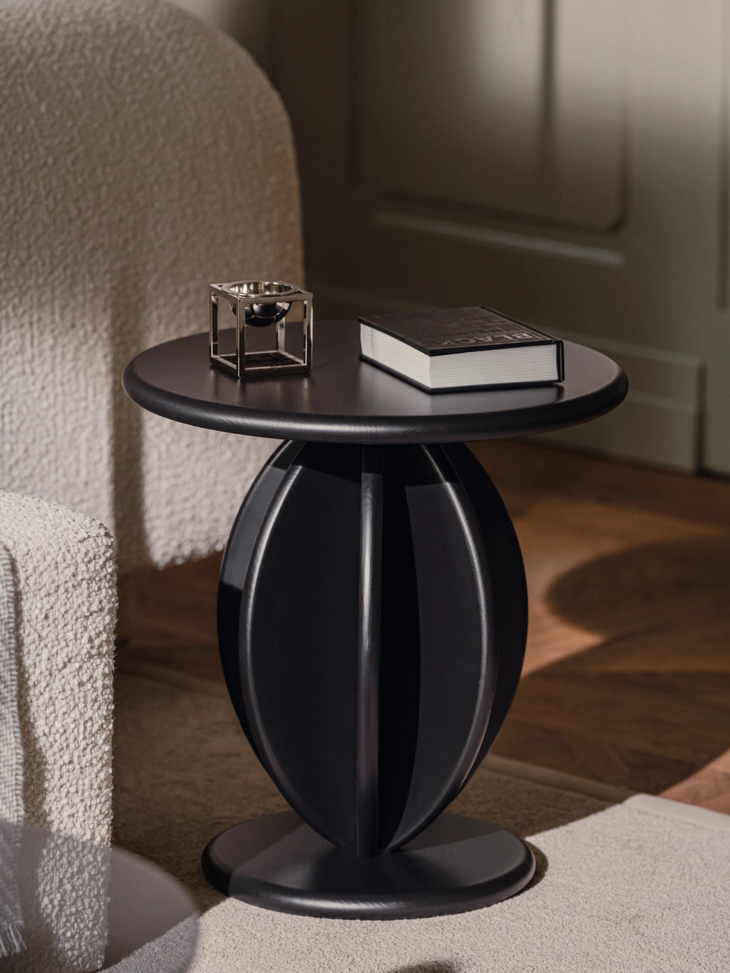TAMO Citrus Side Table in a lifestyle shot. This premium, modern black wooden table features a sculptural, ribbed tangerine-inspired base. Styled with a silver decor piece and book, next to a textured cream armchair. Perfect as a designer living room accent.