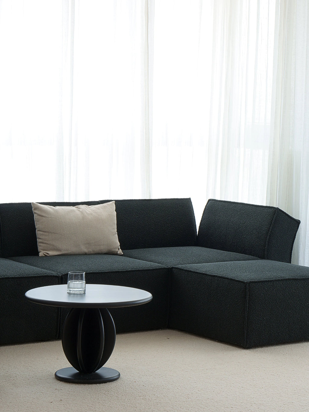 TAMO Citrus Coffee Table, a luxury matte black wood table with a round top and spherical slatted base, styled next to a sofa with a glass of water. A premium accent for contemporary living spaces.
