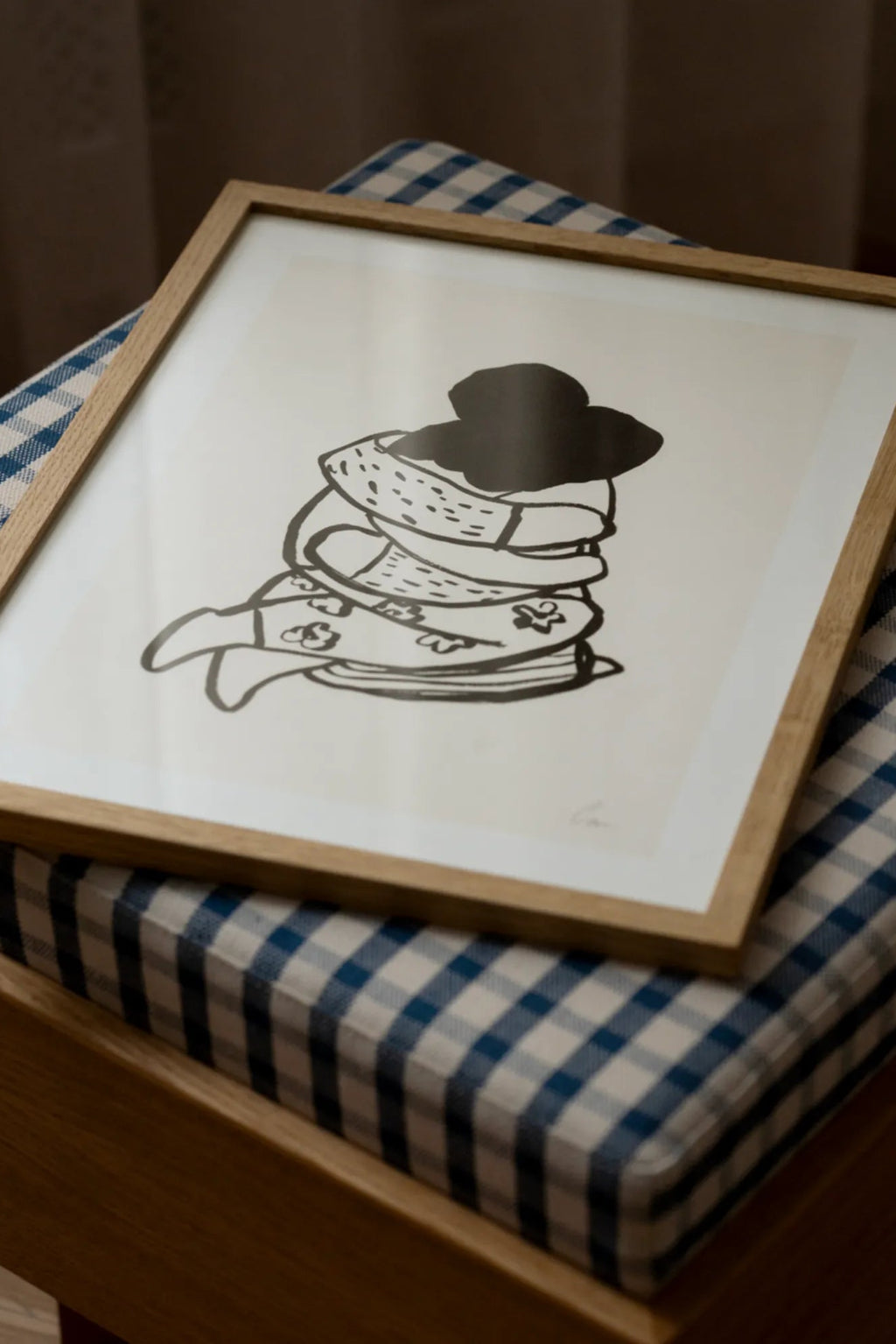 The Poster Club Embrace, a minimalist black illustration art print featuring stacked soft forms topped with a hat, set against a clean white background. This graphic poster is framed in a natural light oak frame with a crisp white mat, enhancing its modern appeal. Ideal as contemporary wall art for Scandinavian bedrooms, minimalist living rooms, or creative gallery walls.