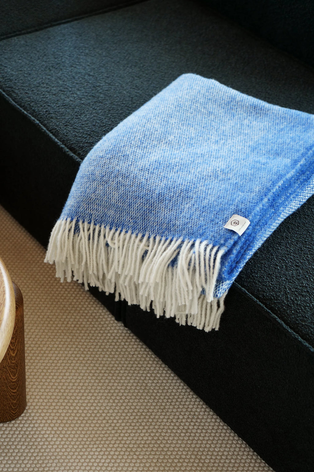 TAMO Meri Blanket Throw, a premium sky blue and white woven natural wool blanket with white fringed edges, folded over a dark textured couch. Ideal for cozy home decor.