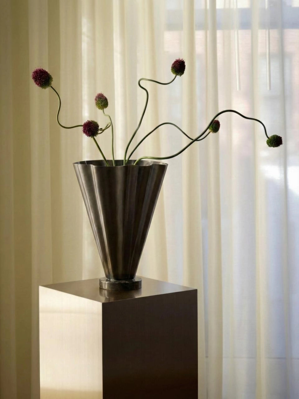 Nordal Coll Vase, a striking modern metal vase with a dark bronze or brushed black finish and a distinctive fluted, conical silhouette. Its elegant design features a wide, flared top with a subtly wavy rim and a sturdy base, perfect for showcasing sculptural botanicals or dried arrangements. An ideal contemporary decorative vase for minimalist living rooms, modern entryways, or sophisticated shelf styling.