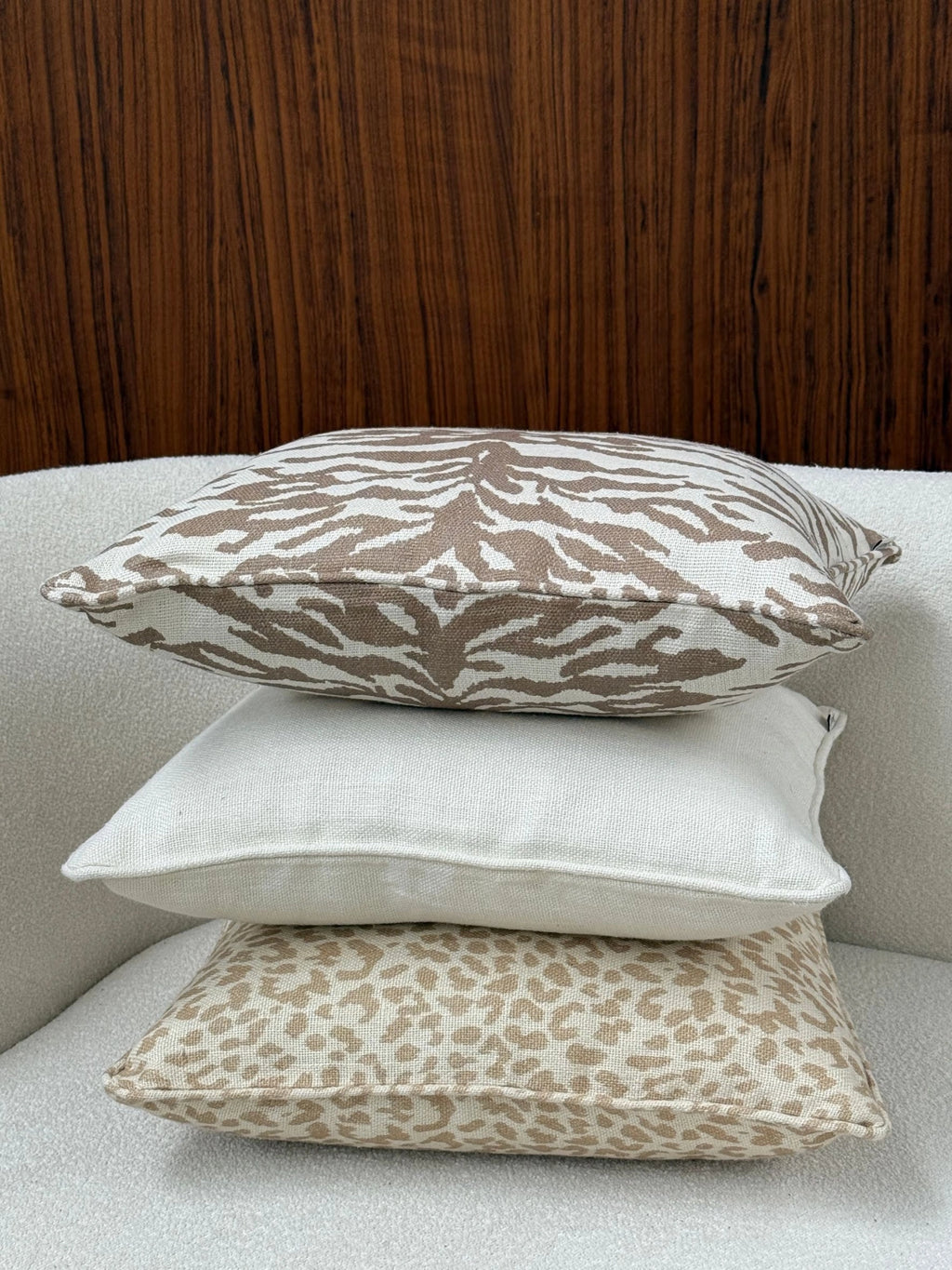 Tinted Objects Norell Throw Pillow, a stack of three elegant decorative cushions on a textured white sofa. The top pillow features a stylish brown zebra print on a cream background, the middle is a plain cream linen, and the bottom has a chic tan leopard print. These versatile cushions are perfect for adding a touch of modern luxury and comfort to any living room or bedroom decor.