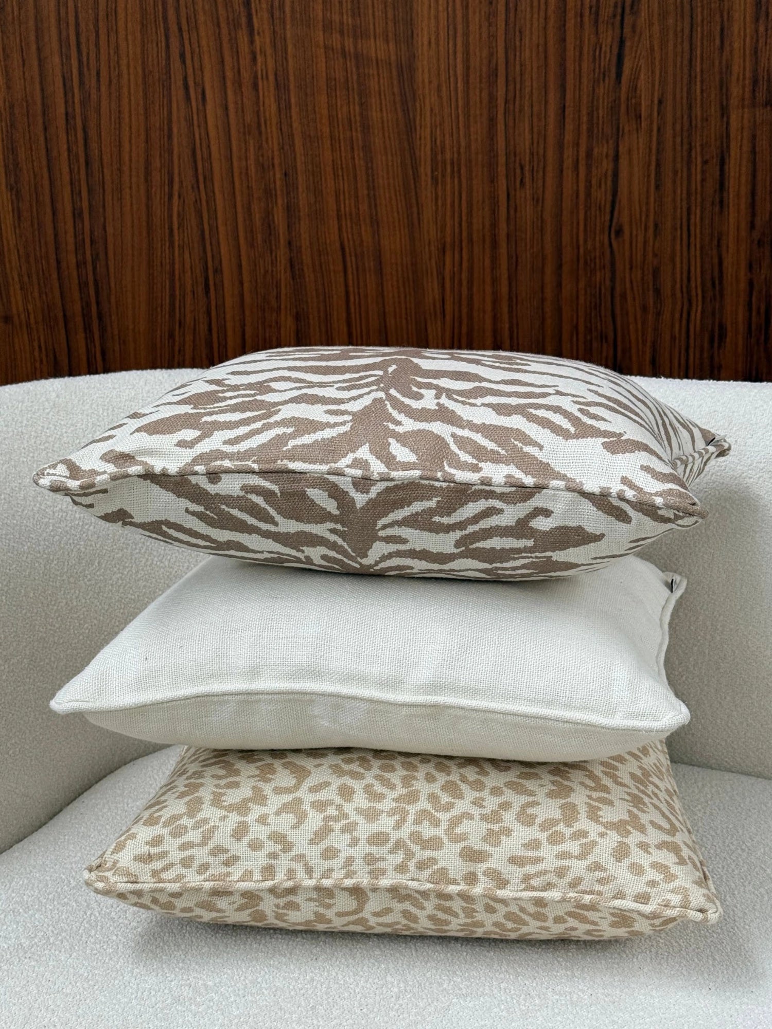 Tinted Objects Norell Throw Pillow, a stack of three elegant decorative cushions on a textured white sofa. The top pillow features a stylish brown zebra print on a cream background, the middle is a plain cream linen, and the bottom has a chic tan leopard print. These versatile cushions are perfect for adding a touch of modern luxury and comfort to any living room or bedroom decor.