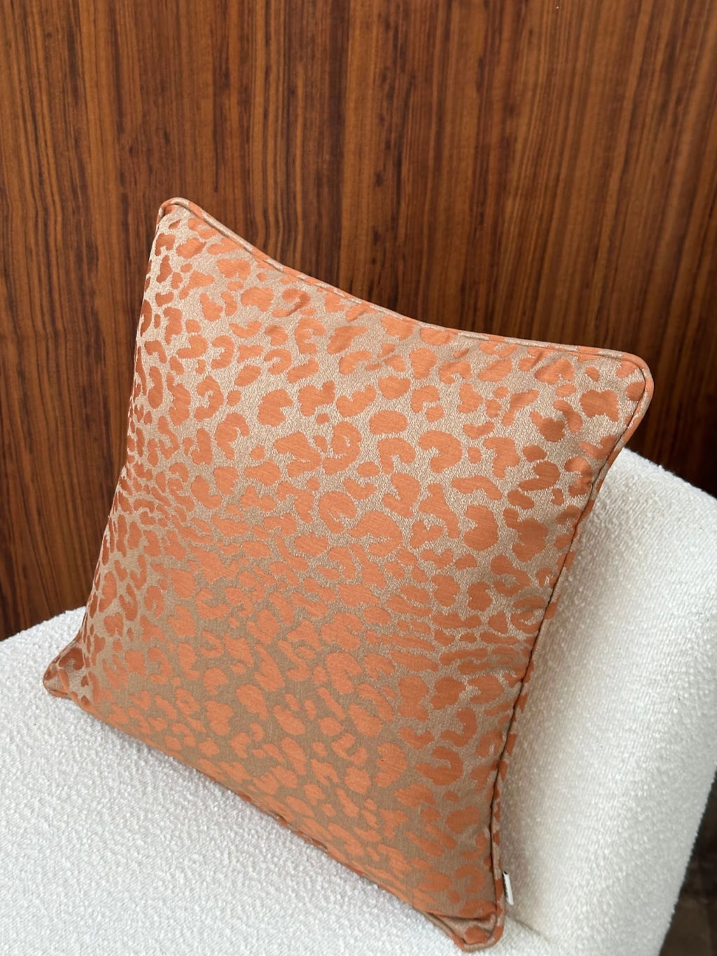 Tinted Objects Vildklint Throw Pillow, a luxurious square cushion featuring a vibrant orange and tan leopard print pattern with a subtle shimmer, resting on a textured white boucle chair. This stylish accent pillow is perfect for adding a touch of modern elegance to any living room or bedroom decor.