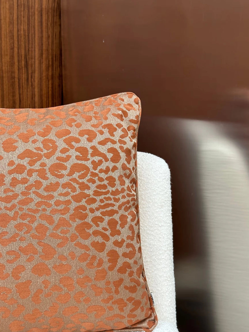 Tinted Objects Vildklint Throw Pillow, a luxurious cushion featuring a raised orange cheetah print on a shimmering gold background, resting against a textured white chair and a rich wooden panel. This stylish accent pillow is perfect for adding a touch of modern elegance and comfort to any living space or bedroom.