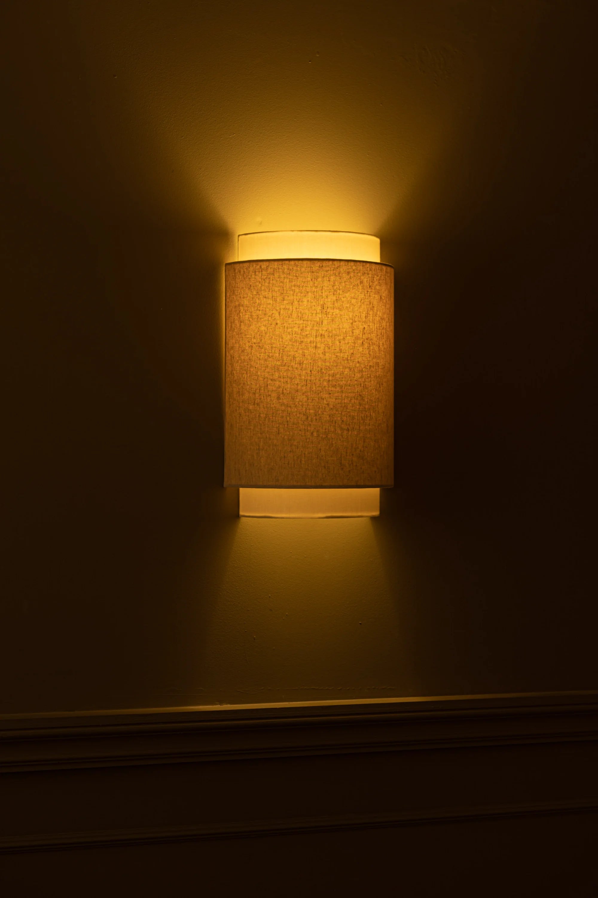 Wall-mounted lamp with a textured shade casting a warm glow on a dark wall.