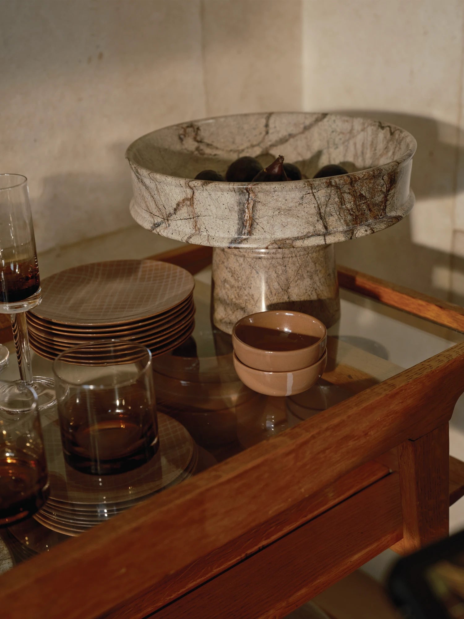 A Dune Raised Bowl made of light brown veined marble containing dark figs is positioned on the glass top of a wooden bar or serving cart. A stack of brown grid-patterned plates, amber-tinted drinking glasses, and small terracotta bowls are also visible on the cart, suggesting a formal serving setting.