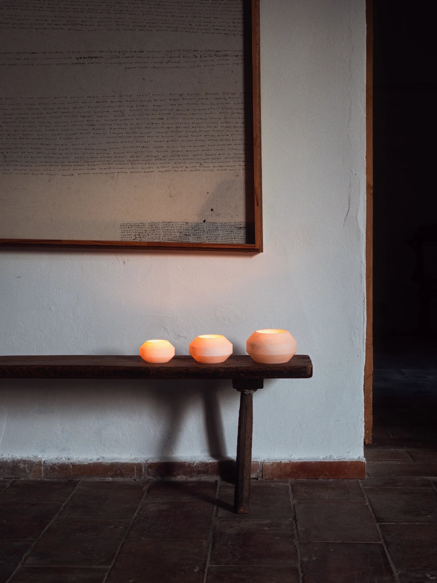 Three faceted alabaster tealight holders of varying sizes are positioned on a dark, rustic wooden bench. All three holders are lit from within, emitting a soft, warm, orange glow against the plain white wall behind them. Above the bench, a large, framed piece of historical or handwritten text hangs on the wall. The floor is tiled with reddish-brown terracotta or brick.