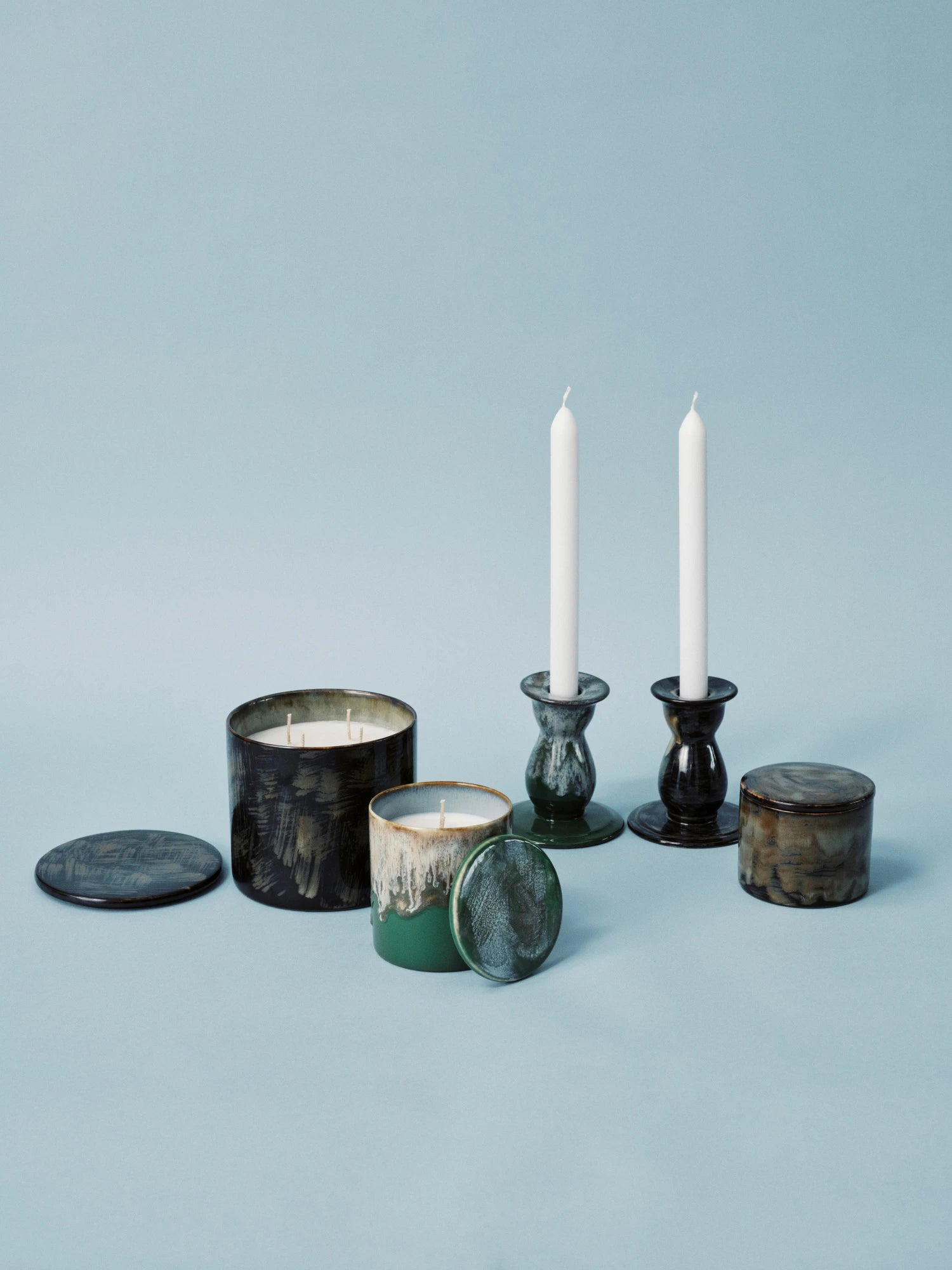 A set of home decor items displayed on a light blue surface. The items include: two Terres De Rêves Ceramic Candle Holders holding unlit white taper candles (one in a glossy dark green/black glaze and one in a mottled green/blue glaze); a large, three-wick candle in a dark, glossy, mottled green/black ceramic container; a smaller, single-wick candle in a green and white mottled ceramic container with its lid resting beside it; and a small, lidded storage jar in a dark brown/green mottled ceramic glaze.