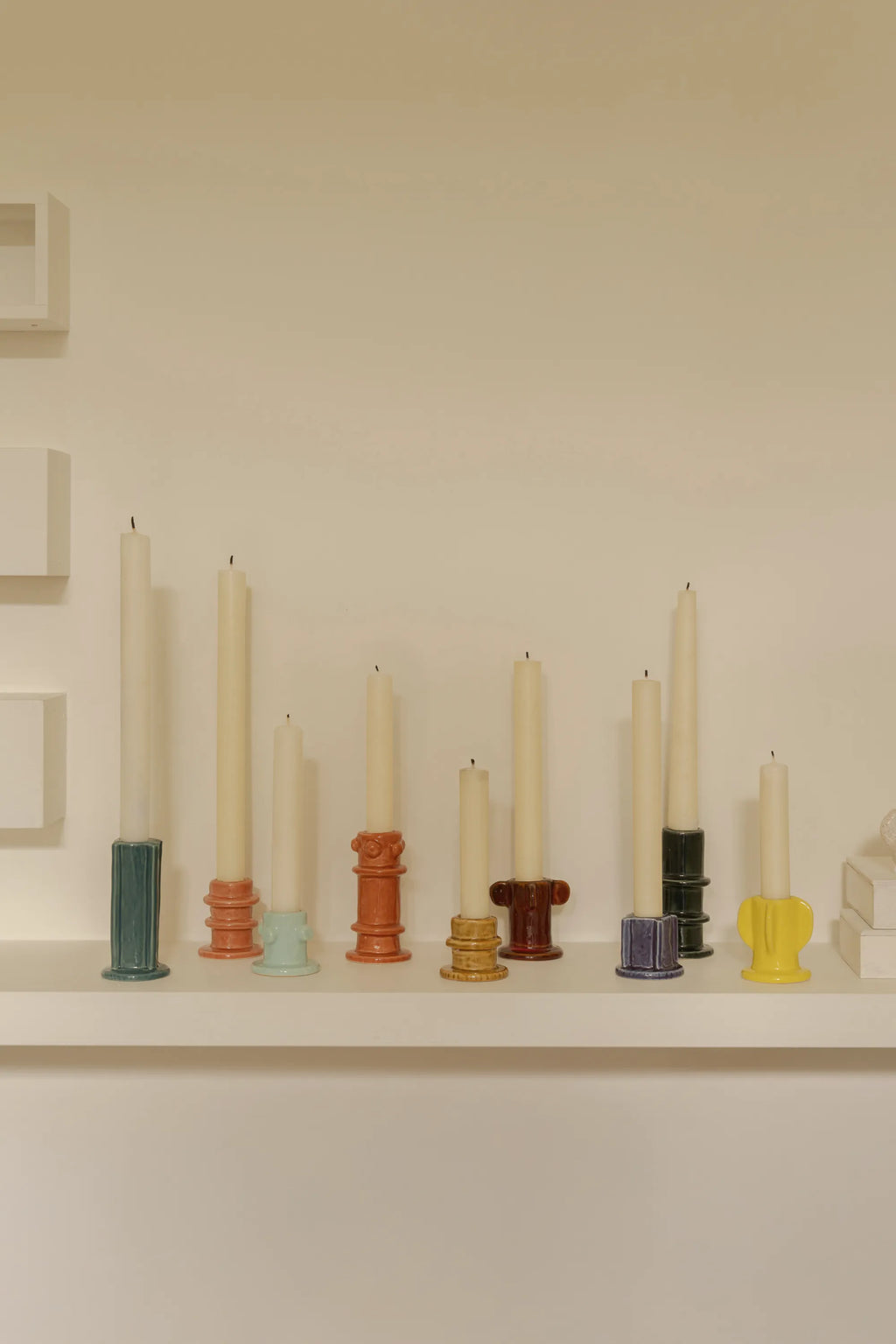 Serax Glazed Molly Candle Holder 01 - Set of 3, featuring a collection of eight unique ceramic pillar candle holders in various vibrant colors including teal, salmon, light blue, terracotta, mustard yellow, dark brown, purple, and dark green, each with a glossy glazed finish and holding a cream-colored pillar candle. These stylish, modern candle holders are perfect for adding a decorative touch to any living space or dining table, ideal for contemporary home decor enthusiasts.