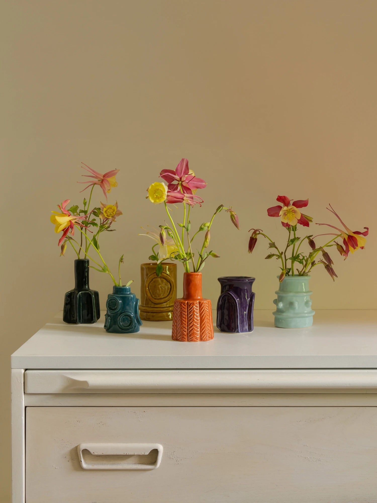 The Molly Vase - Set of 6 is displayed on a rustic white dresser with a single drawer and handle. The six small ceramic vases in glossy black, teal/blue, mustard yellow, bright orange, deep purple, and aqua/mint each hold delicate sprigs of columbine flowers in shades of yellow, pink, and red. The scene is warmly lit against a pale tan wall.