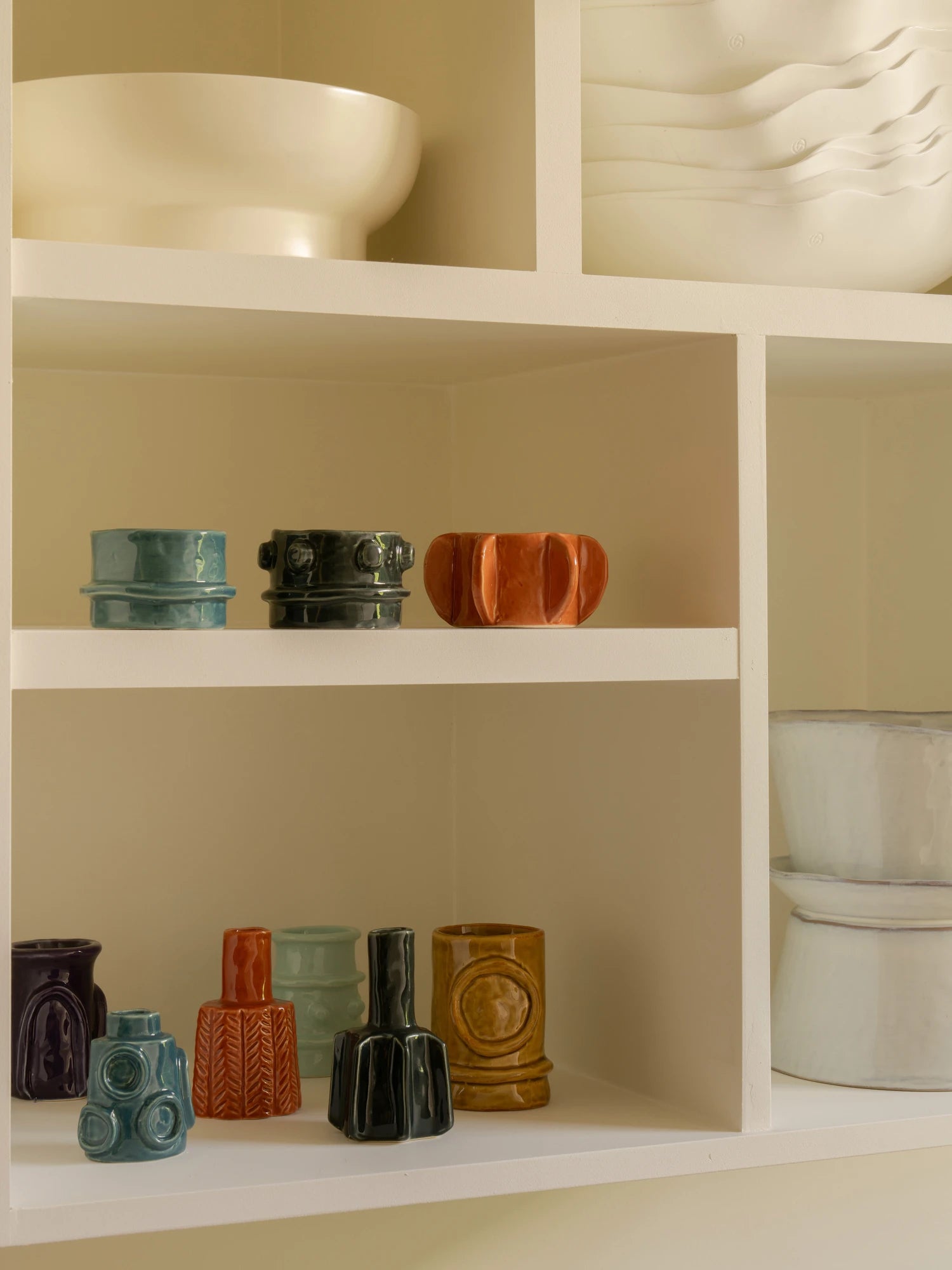 A lifestyle image of the Molly Vase - Set of 6 displayed on light cream-colored shelving units. The set includes six small, unique ceramic vases in various shapes and glazes: a squat teal/blue cylindrical vase, a squat glossy black cylindrical vase, a squat ribbed terracotta/orange vase, a short purple vase, a tall terracotta/orange vase with a zig-zag texture, a tall aqua/mint ribbed vase, a tall glossy black vase with vertical ridges, and a tall mustard yellow vase with a circular relief pattern.