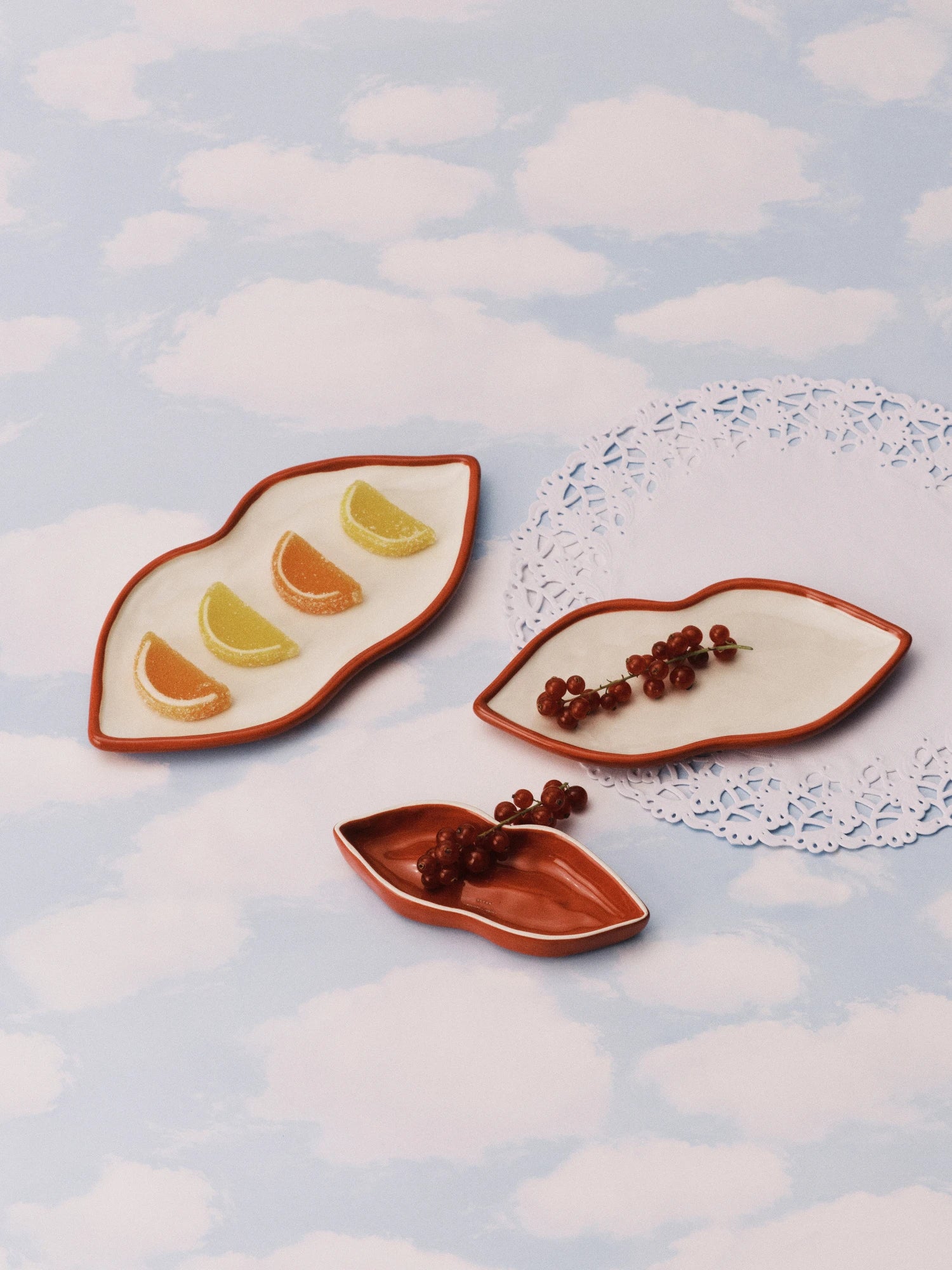A still-life photo showing the Les Objets Mouleversants Serving Dish Set, which includes three ceramic dishes shaped like stylized lips. Two of the dishes are flat with white interiors and burnt-orange rims: the largest holds orange and lemon slice-shaped jelly candies, and the medium one holds a small cluster of red currants. The smallest dish is a deep, solid burnt-orange color and holds a few red currants. The dishes are arranged on a surface decorated with a light blue and white cloud pattern, with a w