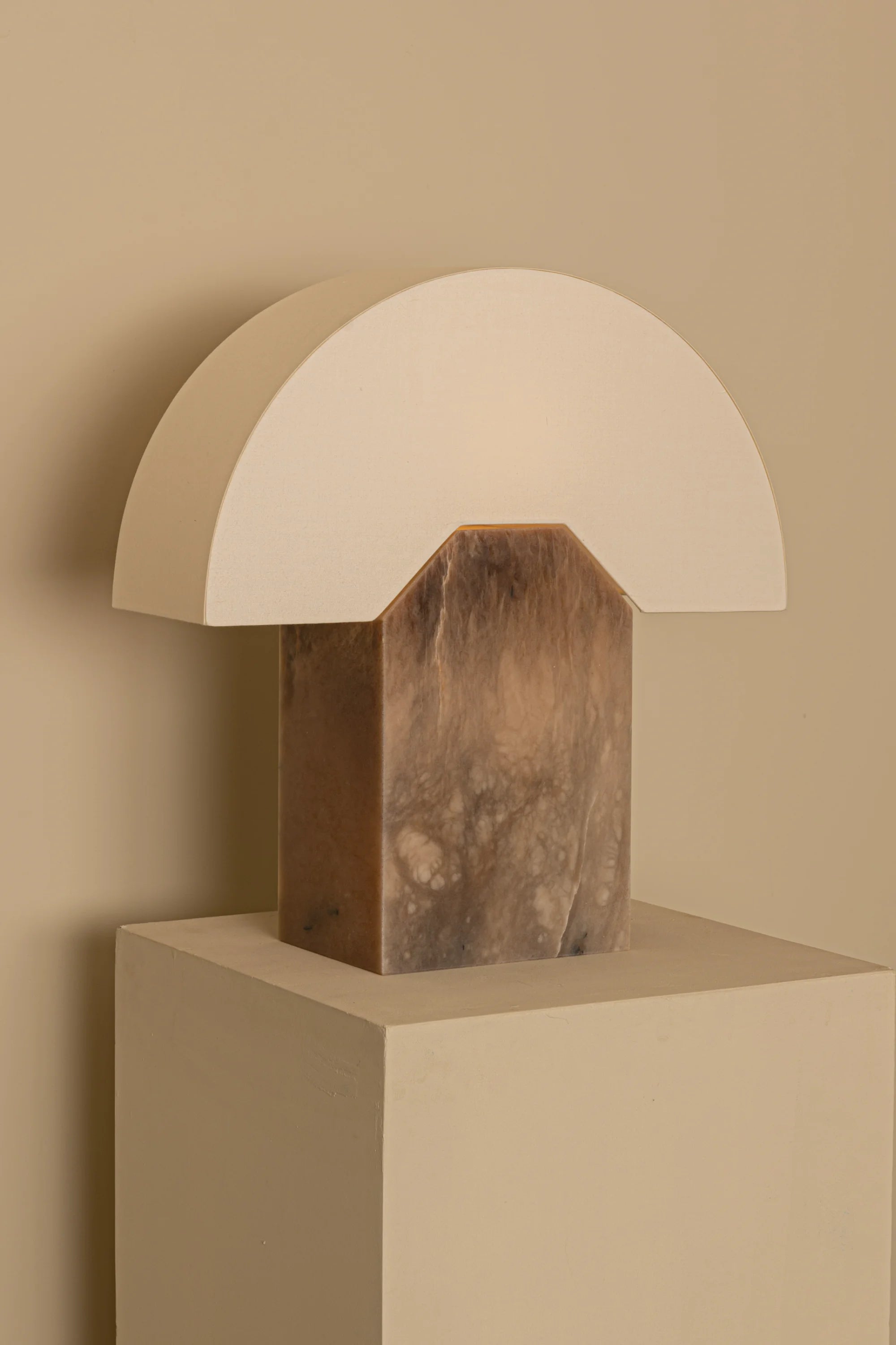 Table lamp with wooden base and white arch-shaped shade on a beige pedestal against a beige wall.
