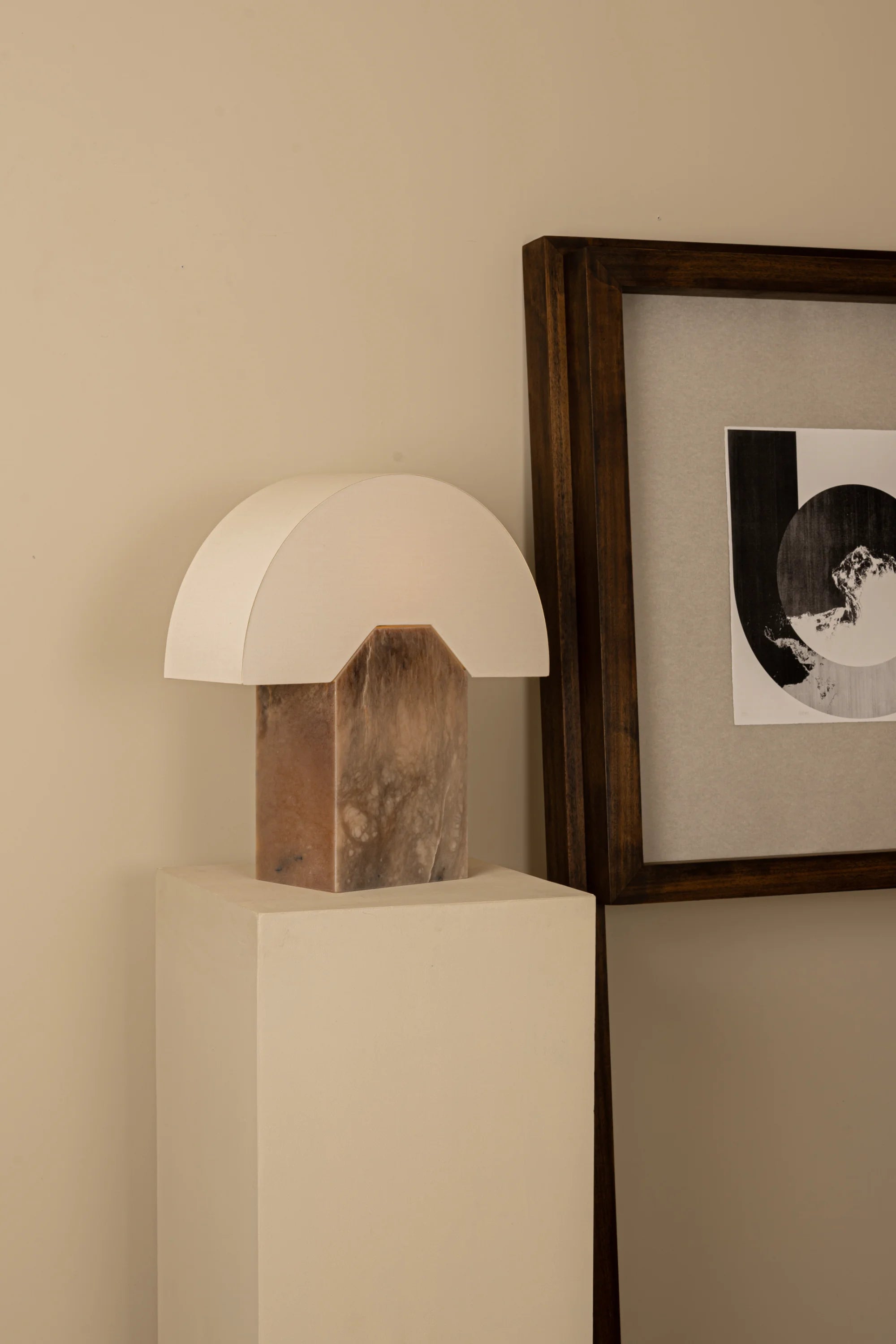 Table lamp with wooden base and white shade on a pedestal against a beige wall with a framed picture.
