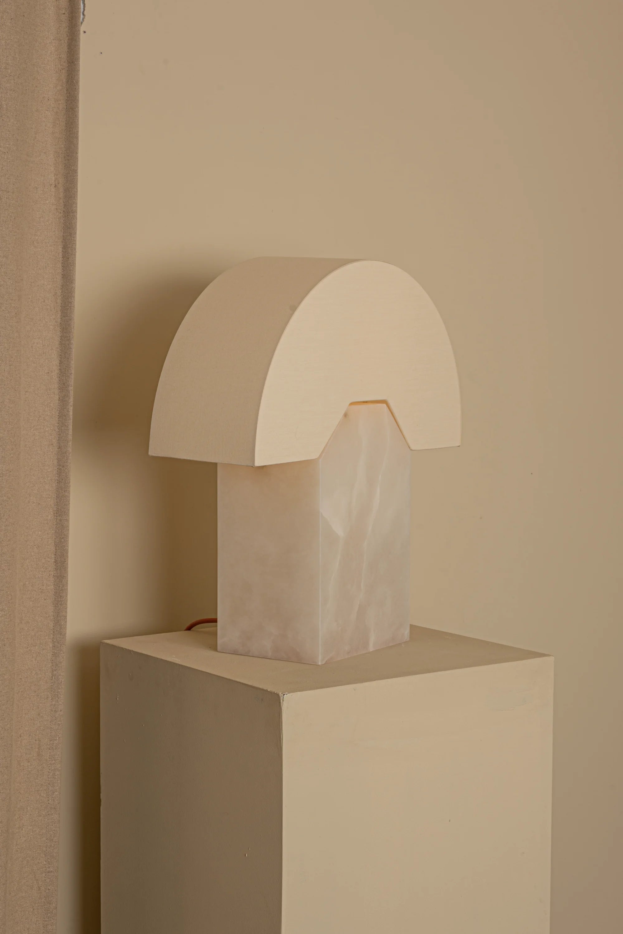 Minimalist table lamp with a alabaster base on a beige pedestal against a beige wall.