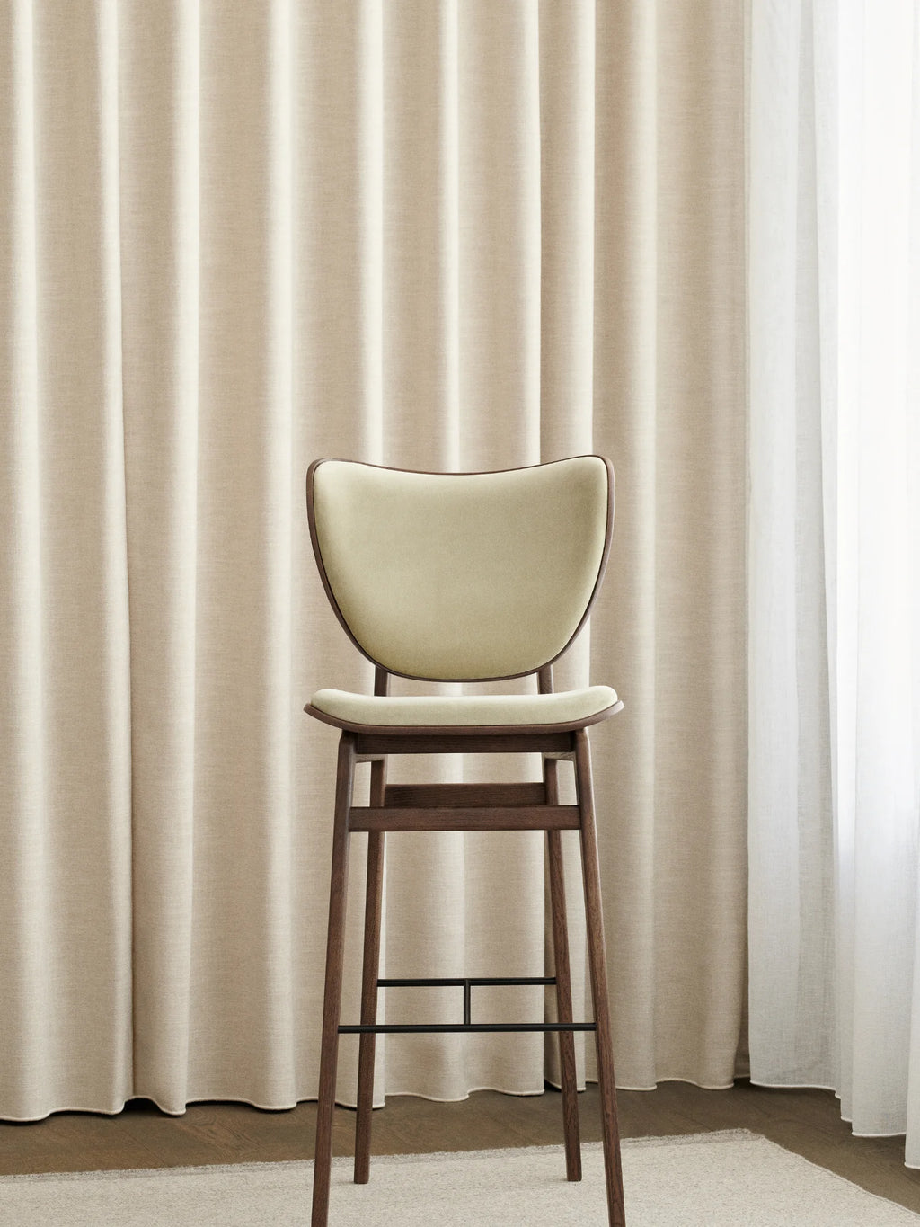 NORR11 Elephant Bar Chair featuring a dark brown wooden frame, curved cream upholstered seat and backrest, and black metal footrest, shown against light curtains. A premium, comfortable bar seating option.