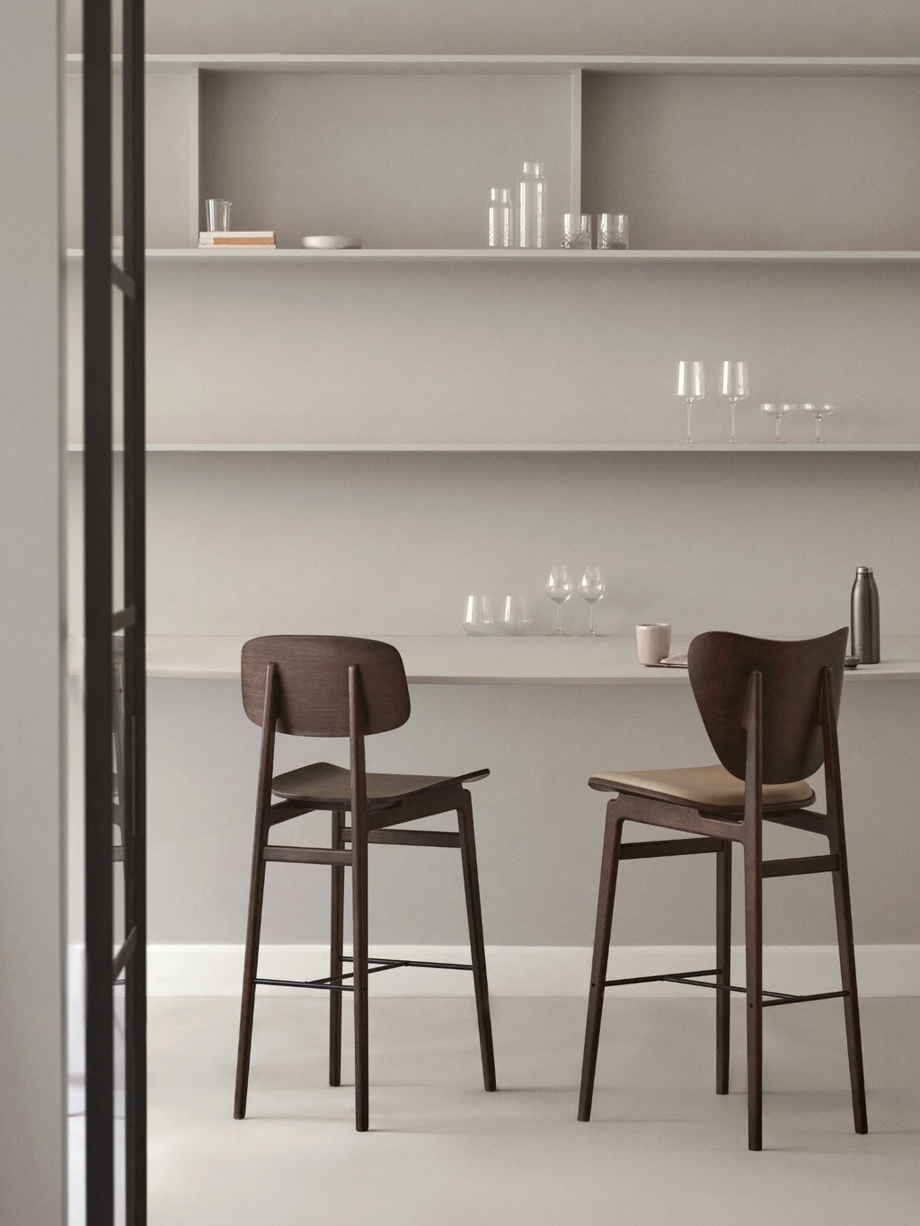 NORR11 Elephant Bar Chair set in a sophisticated kitchen. Two premium dark walnut wood bar stools with curved backs and natural leather-padded seats are positioned against a minimalist light grey wall with shelves of glassware. Ideal for modern dining.