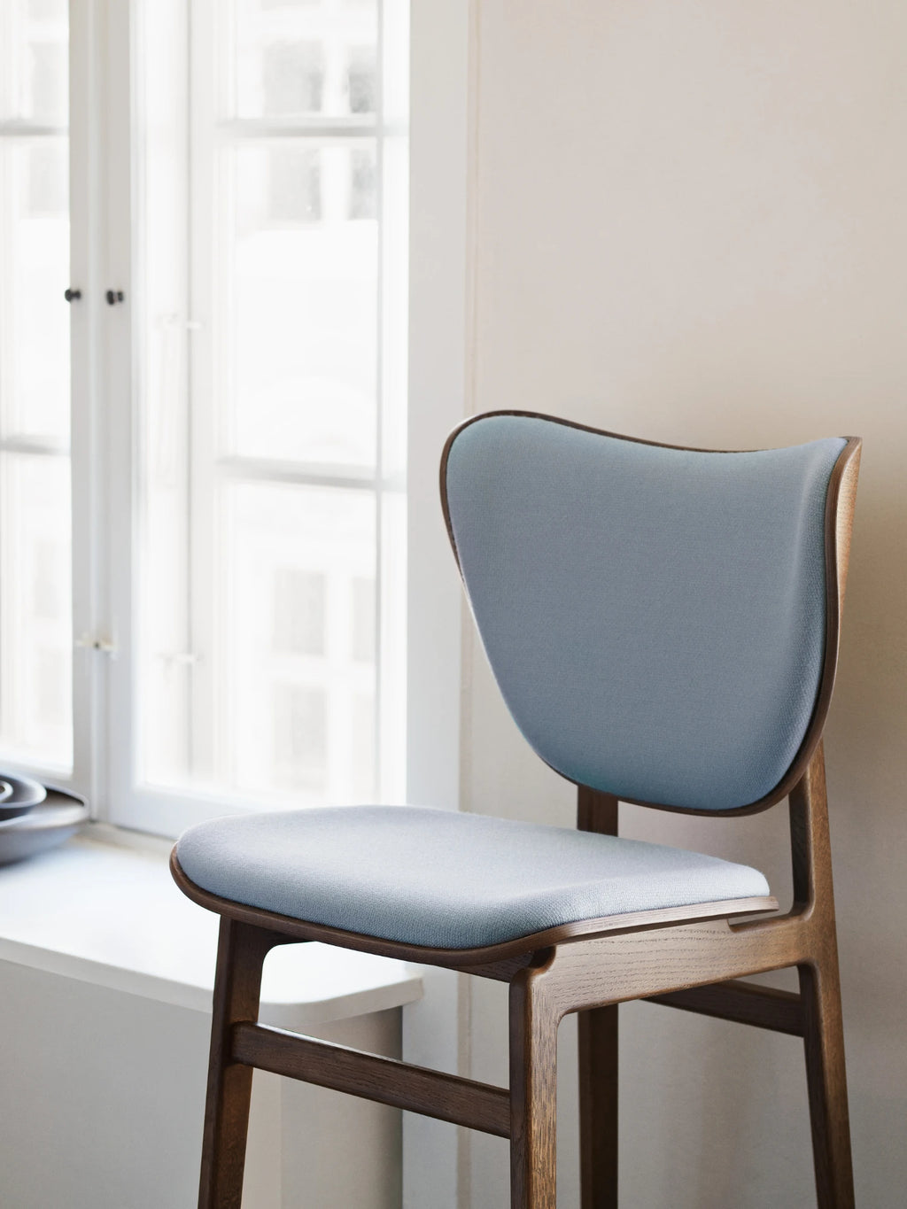 NORR11 Elephant Chair, a modern Scandinavian dining chair crafted with a gracefully curved dark oak wood frame featuring a visible grain and soft light blue woven fabric upholstery on its rounded seat and distinctive elephant ear-shaped backrest. This minimalist wood dining chair is perfect for contemporary dining rooms, Nordic-inspired kitchens, or as a stylish accent piece in a modern living space.