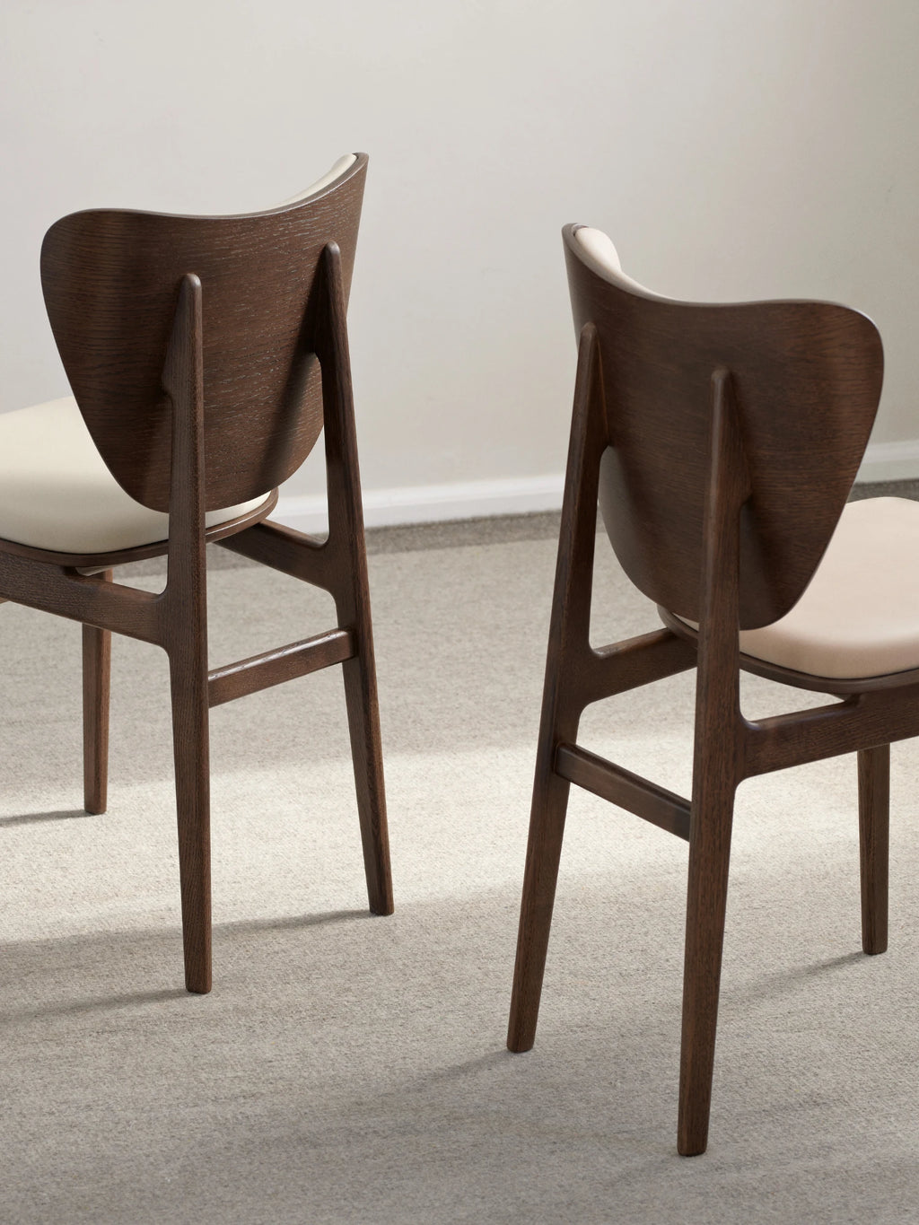 NORR11 Elephant Chair, a distinctive mid-century modern dining chair featuring a dark stained oak wood frame with a gracefully curved, organic backrest and sleek tapered legs. It boasts a comfortable cream-colored upholstered seat, offering a refined blend of Scandinavian design and contemporary elegance. Perfect as a stylish dining room chair or an accent piece in a modern living space.