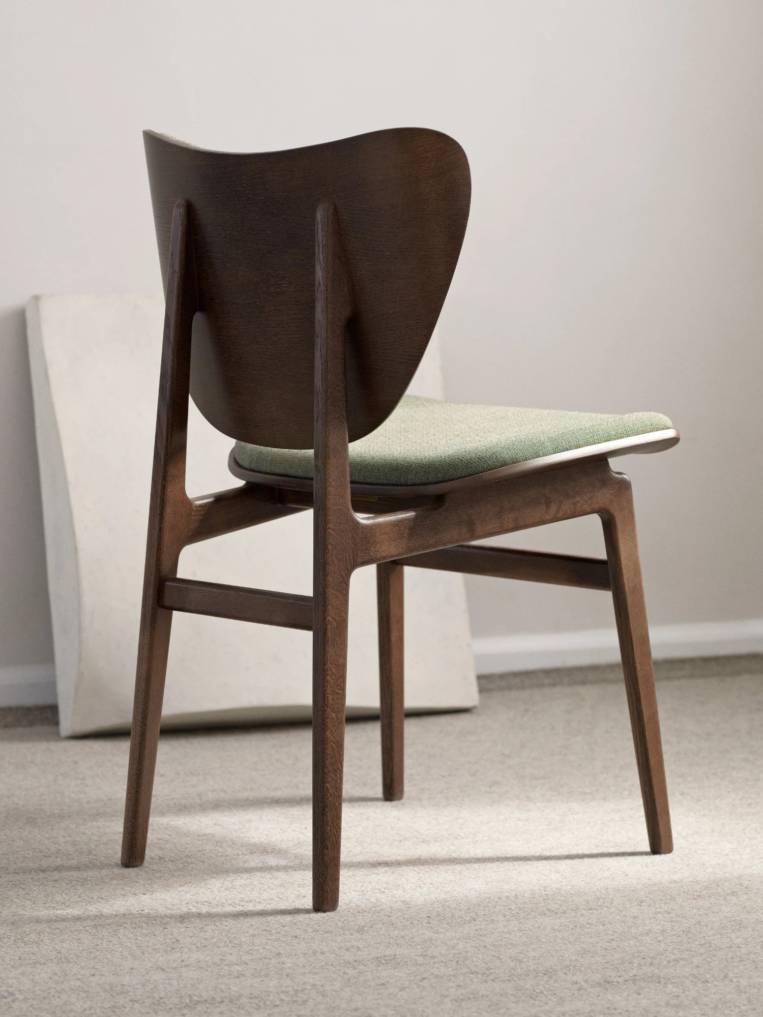 NORR11 Elephant Chair, a mid-century modern dining chair crafted from dark stained solid oak with a distinctive curved organic backrest and a comfortable upholstered seat in a light sage green textured fabric. This contemporary wood chair features sleek, tapered legs, perfect as a stylish dining room chair or an elegant accent chair for Scandinavian-inspired interiors.