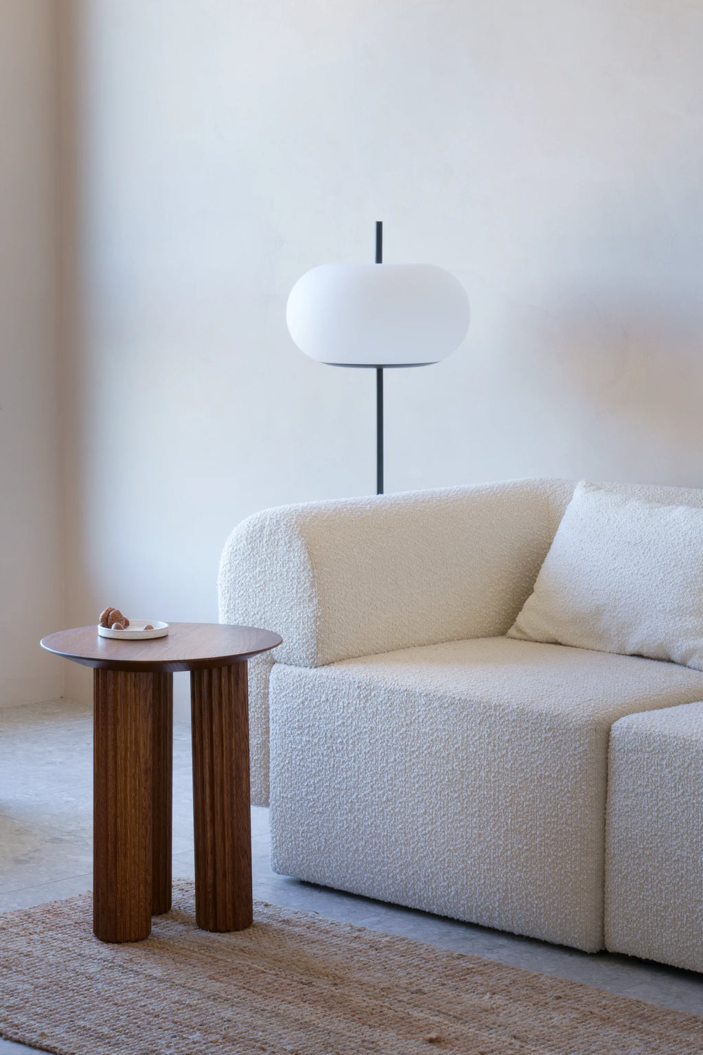 TAMO Filar Side Table, a round dark brown wooden table with three fluted, ridged legs, styled with a small white dish and nuts on top, beside a cream bouclé sofa. This luxury accent piece is ideal for modern living spaces.