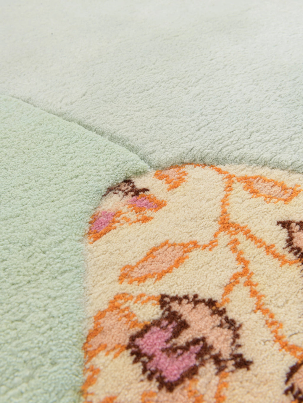 Haus Üger Flora Rug, a luxurious hand-tufted wool area rug showcasing a unique organic shape with a soft, plush pile. It features a modern design combining a solid light sage green section with a cream base intricately patterned with vibrant floral motifs in orange, soft pink, and brown tones. Perfect for adding a playful, bohemian touch to contemporary living rooms, cozy bedrooms, or as a whimsical accent in a nursery or kids' room.