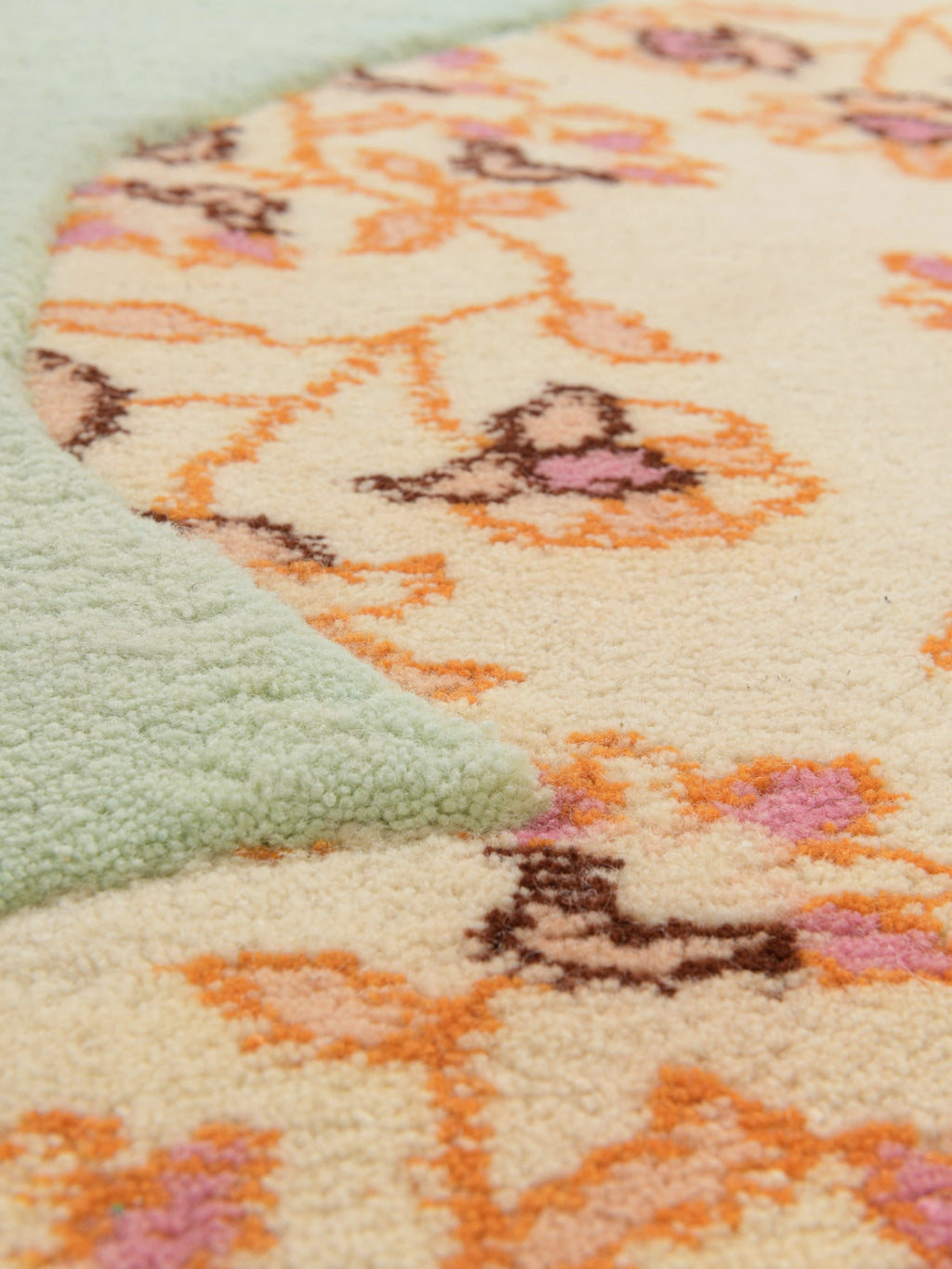 Haus Üger Flora Rug, a whimsical hand-tufted wool area rug featuring a plush cream-colored pile adorned with a vibrant floral pattern in shades of orange, pink, and brown. An organic, curvilinear border in soft mint green adds a playful, contrasting texture and shape. This cozy, retro-inspired accent rug is perfect for adding boho charm to a contemporary living room, bedroom, or nursery decor.