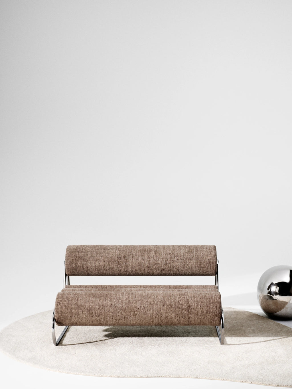 NORR11 Fave Sofa, a modern minimalist lounge chair featuring two horizontal cylindrical cushions upholstered in a warm brown textured fabric, supported by a sleek polished chrome tubular metal frame. This contemporary compact sofa offers a clean, industrial chic aesthetic, making it perfect for stylish living rooms, apartment lounges, or minimalist office spaces.