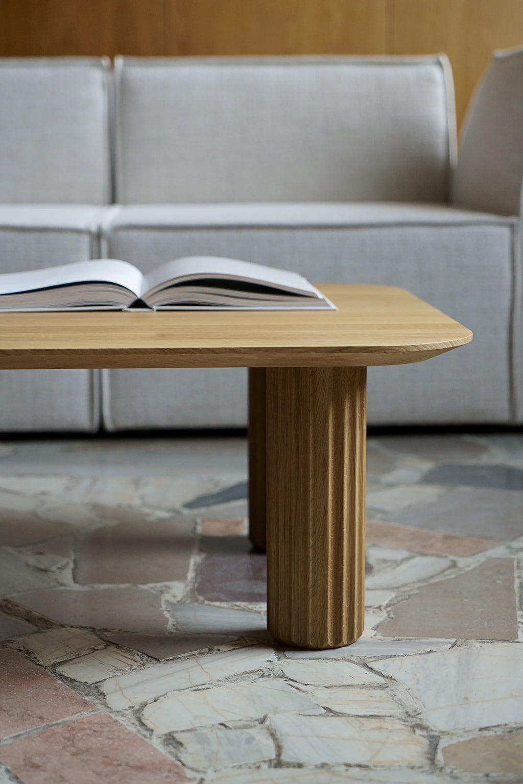 TAMO Filar Coffee Table, a minimalist rectangular solid light oak wood table with two fluted pillar legs, and a book on top, in a contemporary living room setting. A premium statement piece for modern interiors.