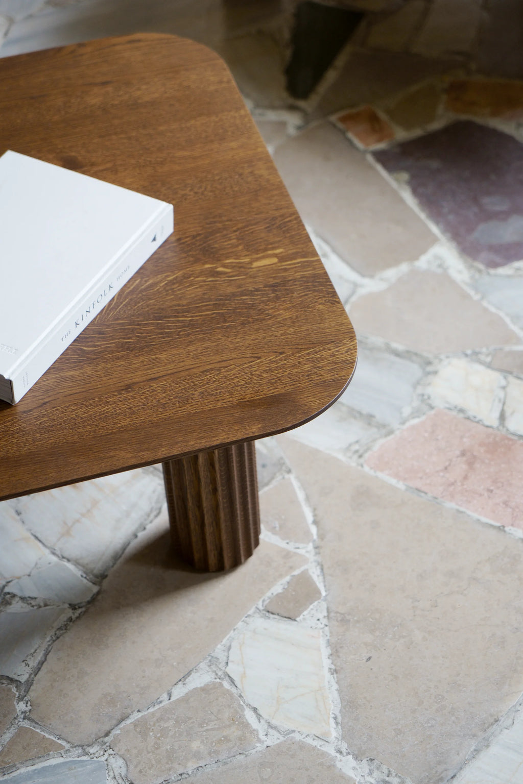 TAMO Filar Coffee Table, a modern, rounded rectangular tabletop of rich brown wood with visible grain texture, atop a fluted base. A white book sits on the surface. Ideal for contemporary living spaces.