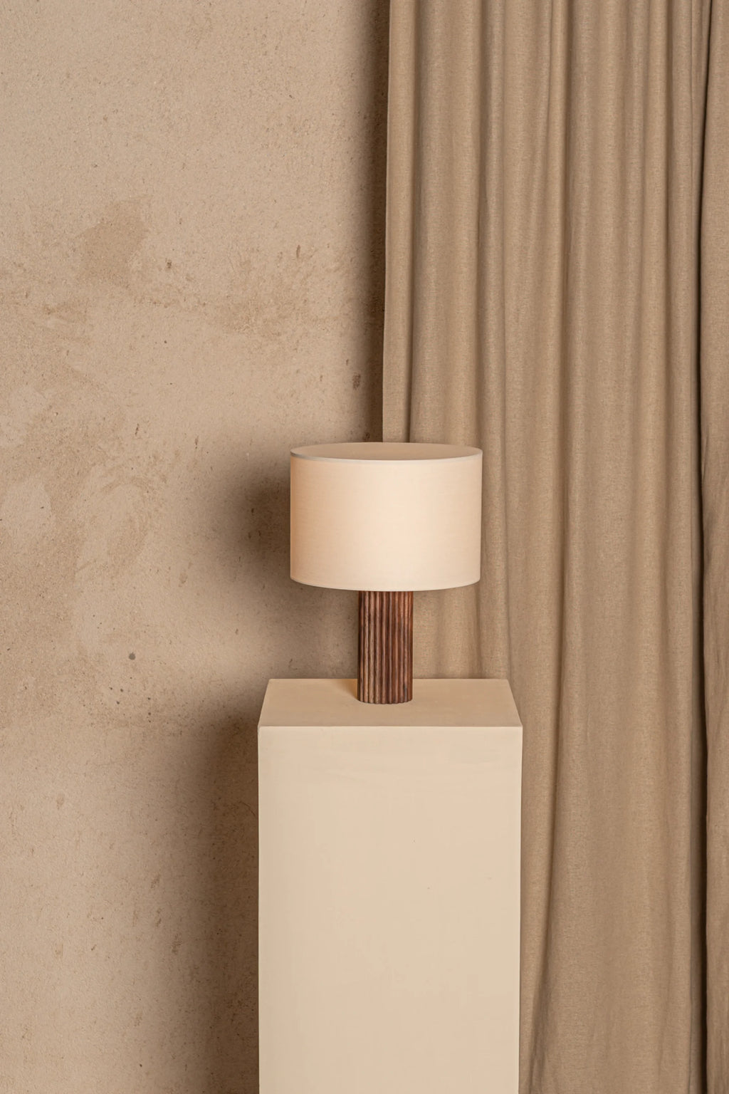 Table lamp with wooden base and beige shade on a white pedestal against a beige wall and curtain.